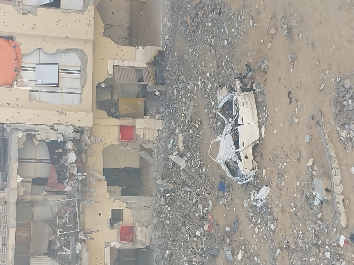 The image displays a scene of widespread destruction in Khan Yunis, Palestinian Territory. In the background, a multi-story building with a light-colored facade shows extensive structural damage. Large sections of its exterior walls and interior floors are missing, exposing destroyed rooms filled with debris, twisted metal, and broken furniture. The remaining facade is heavily pockmarked with numerous impact holes. On the ground floor, gaping holes reveal internal spaces; a red cabinet-like object is visible within the left opening, and a yellow cabinet with a detached louvered door is present within the right opening. The ground leading up to the building is covered in a vast expanse of concrete rubble, fragmented building materials, and dirt. In the mid-ground, a white passenger car is severely compressed and mangled, its body panels twisted and collapsed, rendering it unrecognizable. No individuals are present in the scene.