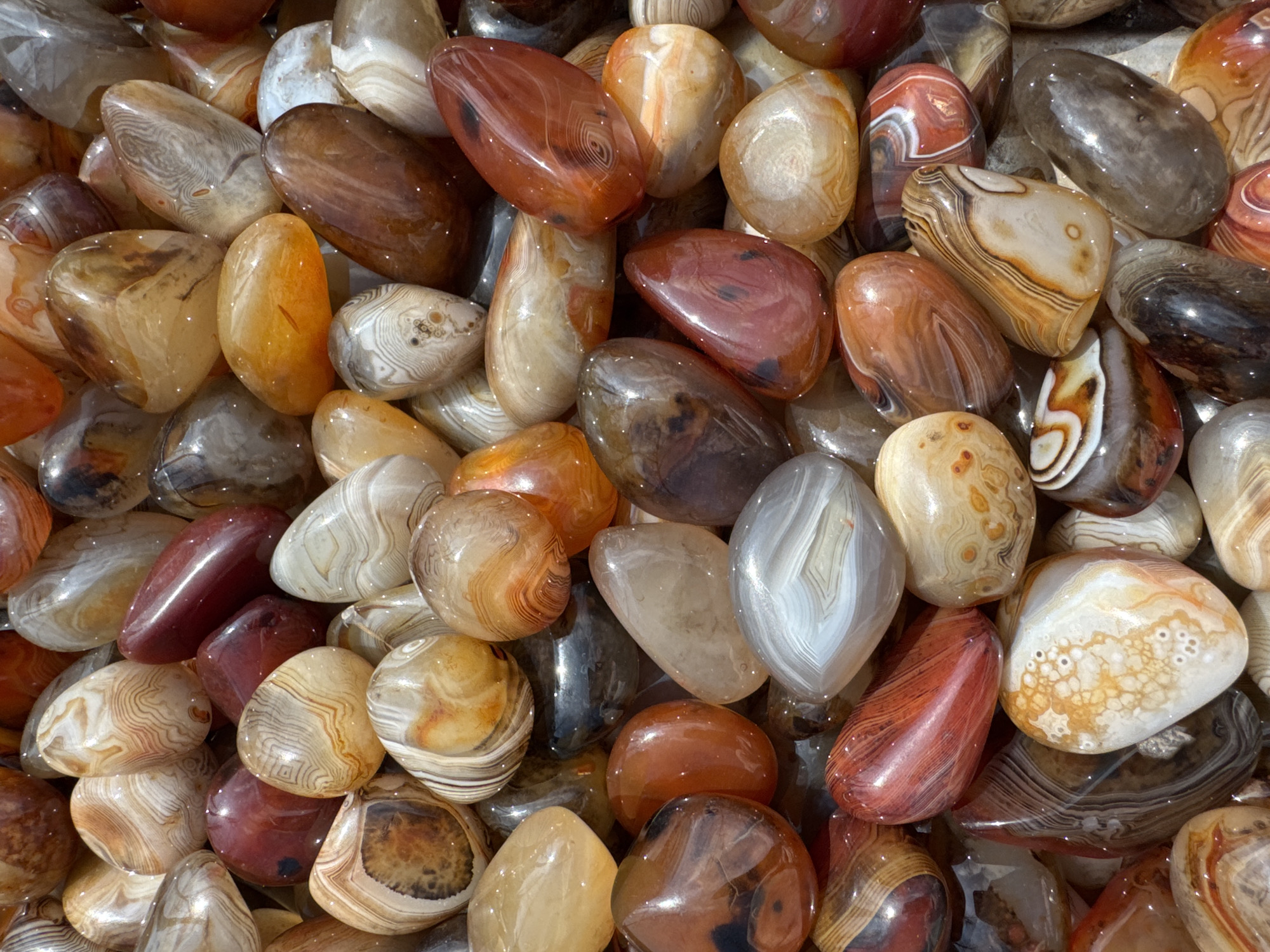 The image presents a dense, top-down view of a large collection of tumbled agate stones. The stones vary in size and irregular, rounded shapes, filling the entire frame. Their polished surfaces exhibit a glossy finish, reflecting ambient light. The coloration includes a spectrum of natural tones such as reddish-brown, orange, cream, white, gray, and translucent hues. Many stones display characteristic banding patterns, concentric rings, or mottled textures. No individuals, subjects, or actions are depicted. The objects are static. This scene is located in Wulingyuan, China.
