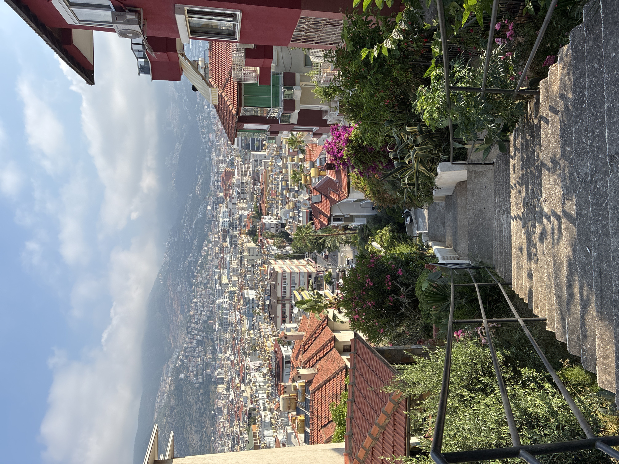 The image captures a view from a set of concrete outdoor stairs descending a hillside in Alanya, Turkey. The stairs are flanked by metal railings and lush greenery, including flowering bushes with pink blossoms and large-leafed plants. In the foreground, the sun casts distinct shadows on the steps. Beyond the immediate stairs, a dense cityscape unfolds, characterized by numerous buildings with red-tiled roofs and a variety of architectural styles. The city extends towards a hazy mountain range in the background, which is partially obscured by clouds. The sky is blue with scattered white clouds. The scene suggests a residential area on a slope overlooking the main part of the city.