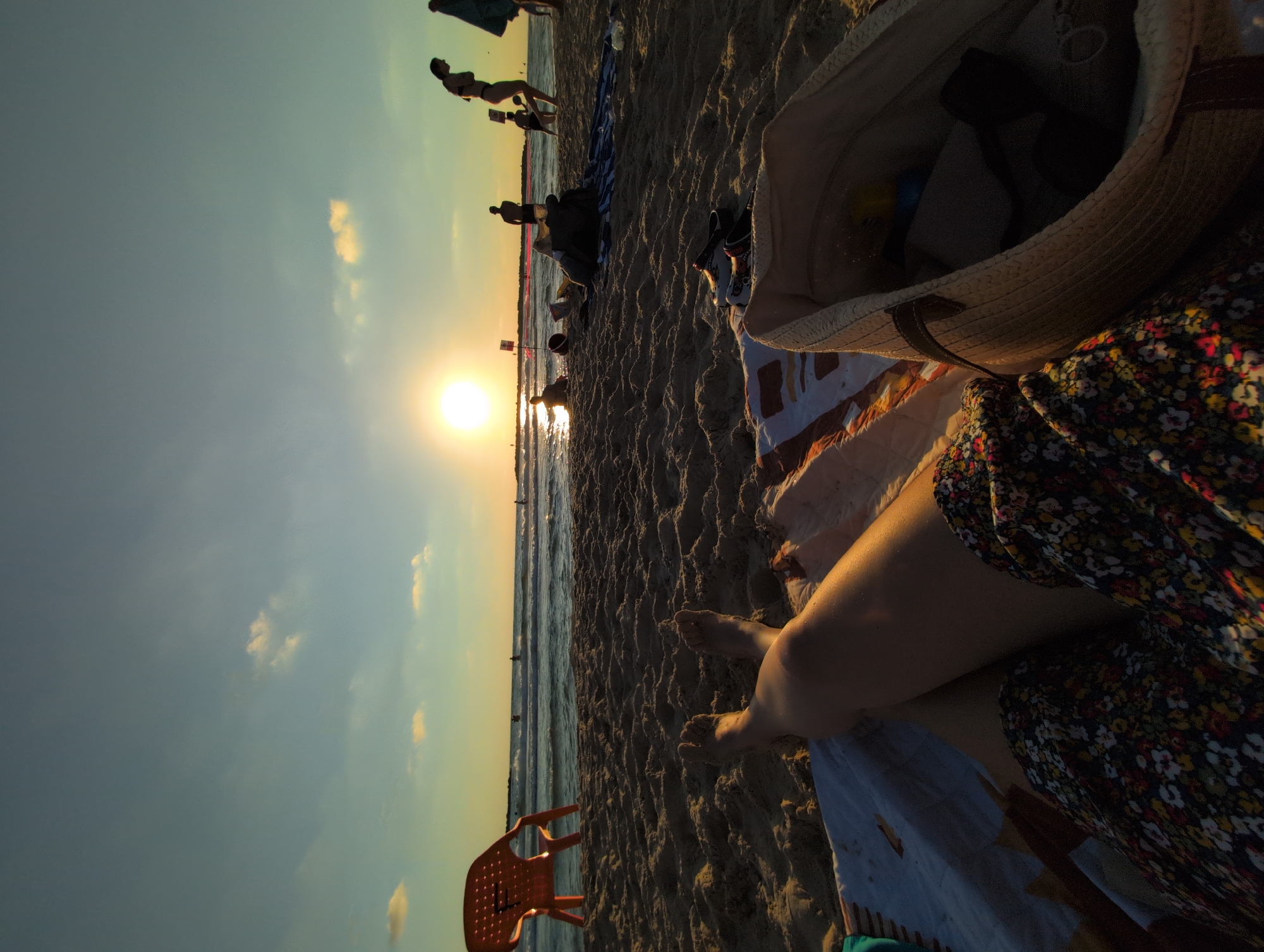 A low-angle shot depicts a beach scene at sunset in Tel-Aviv, Israel. The foreground is dominated by the legs and floral-patterned skirt of a person relaxing on a blanket. A straw beach bag sits to the right. The sand is textured with footprints and small ripples. In the midground, other beachgoers are visible, some wading in the shallow water, while others sit on the sand. An orange plastic chair stands to the left. The sea stretches to the horizon, where the sun, a bright orb, is setting, casting a warm glow and reflecting on the water. The sky is a gradient of blue and yellow, with a few scattered clouds. The overall impression is one of a tranquil evening at the beach.
