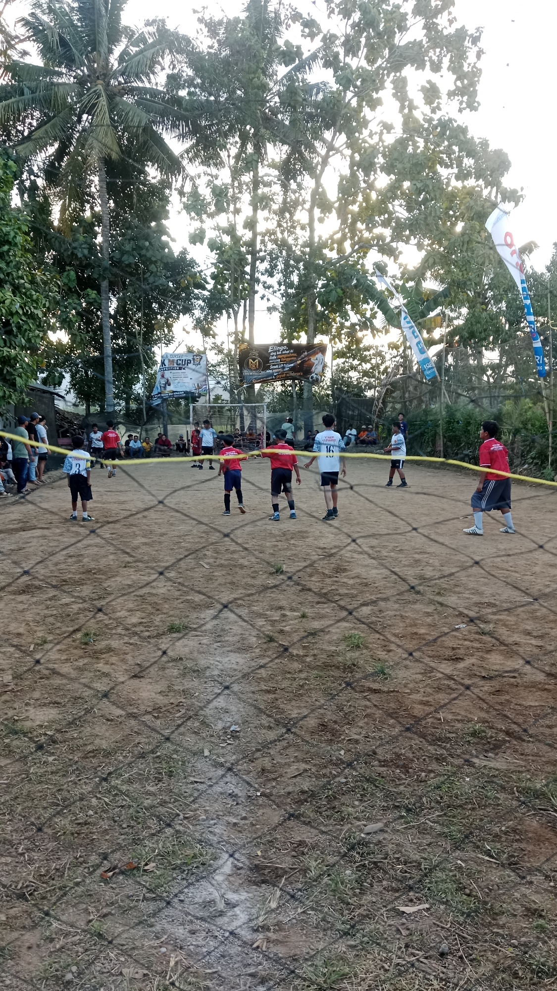 In Karangharjo, Indonesia, an outdoor amateur soccer match is underway on a dirt field. The sun is low in the sky, casting long shadows. A net is strung across the foreground, obscuring some of the action.
Six young players are visible on the field, dressed in red and blue uniforms. Two players in red jerseys and blue shorts are facing each other, one with his arms outstretched, possibly in a defensive or pleading gesture. Another player in a red jersey and dark shorts stands nearby, looking towards the other players. Two players in blue jerseys and black shorts are also on the field, one with a ball at his feet and the other facing away from the camera. A sixth player, in a white jersey with the number "16" and blue shorts, stands further down the field. Spectators are gathered behind a fence on the left side of the field, and some are seated on benches behind the goal on the right.
Two banners are visible in the background, one white with blue text and a logo, and another with a stylized "M" logo and an image of a soccer player. Several tall trees with dense foliage surround the field, and flags are mounted on poles to the right. The ground is uneven and dusty, with patches of dry grass. A white line, likely marking the edge of the field, is visible in the foreground.
