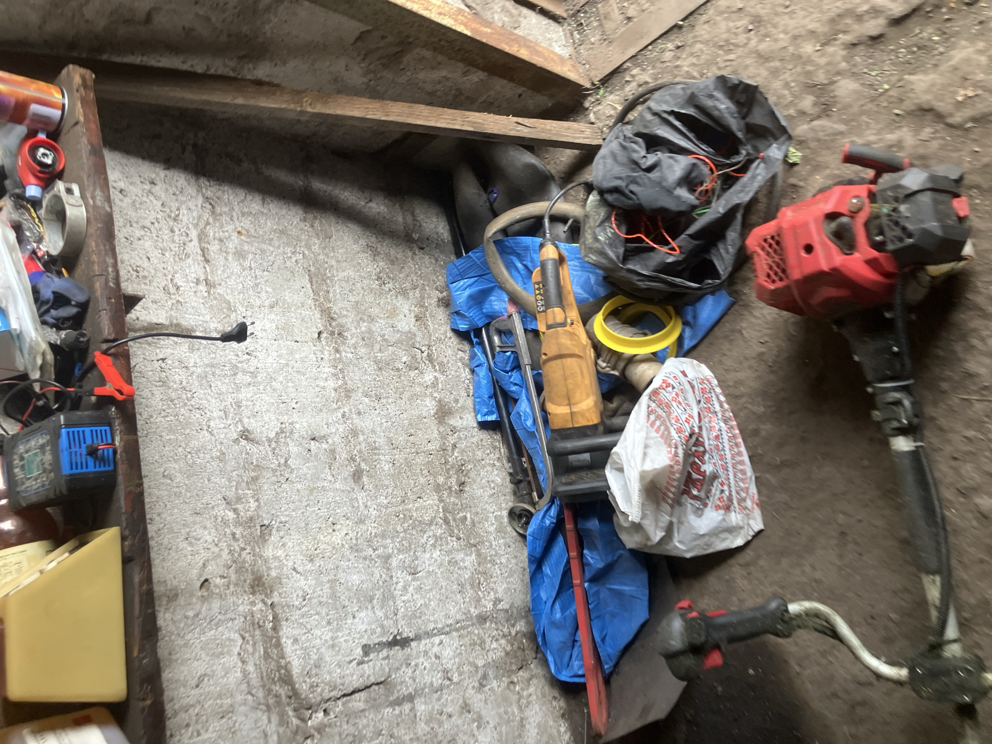 In Vovkivka, Ukraine, a cluttered corner of what appears to be a workshop or storage area is depicted. A wooden shelf, stained and worn, is mounted on a rough, concrete wall. On the shelf sit various items including a yellow wedge-shaped object, a blue and black battery charger with red clamps, a metal clamp, and a can. Below the shelf, a collection of tools and materials are piled on the ground. A bright blue tarp covers some items, beneath which a red bar is visible. A yellow rotary tool with an orange body and handle is prominently placed. Next to it, a white plastic bag with red lettering is partially visible. To the right, a large black plastic bag contains tangled orange and green wires. In the foreground, a red and black string trimmer lies on the dirt floor, its metal shaft curving towards the left. The environment is dusty and appears to be an indoor space with natural lighting casting shadows.