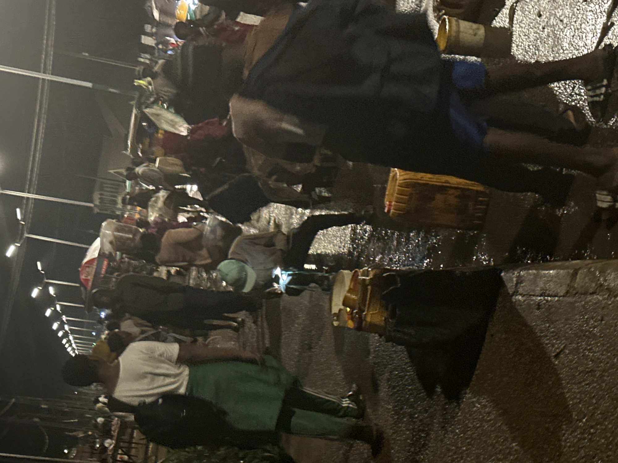 The image depicts a bustling night market scene in Bukuru, Nigeria. In the foreground, two individuals are partially visible, one wearing a white t-shirt and green skirt and the other in dark clothing. Several large yellow barrels and a smaller white one are present, some containing liquid. A person in a light-colored shirt and green cap is seen examining a mobile phone illuminated by its screen. In the background, a crowd of people navigates through the market, with streetlights illuminating the area. Stalls with goods are set up, and umbrellas, one red and white, are visible. The ground appears wet, reflecting the overhead lighting, suggesting recent rain or the presence of water in the market. The overall atmosphere is one of activity and commerce under the night sky.