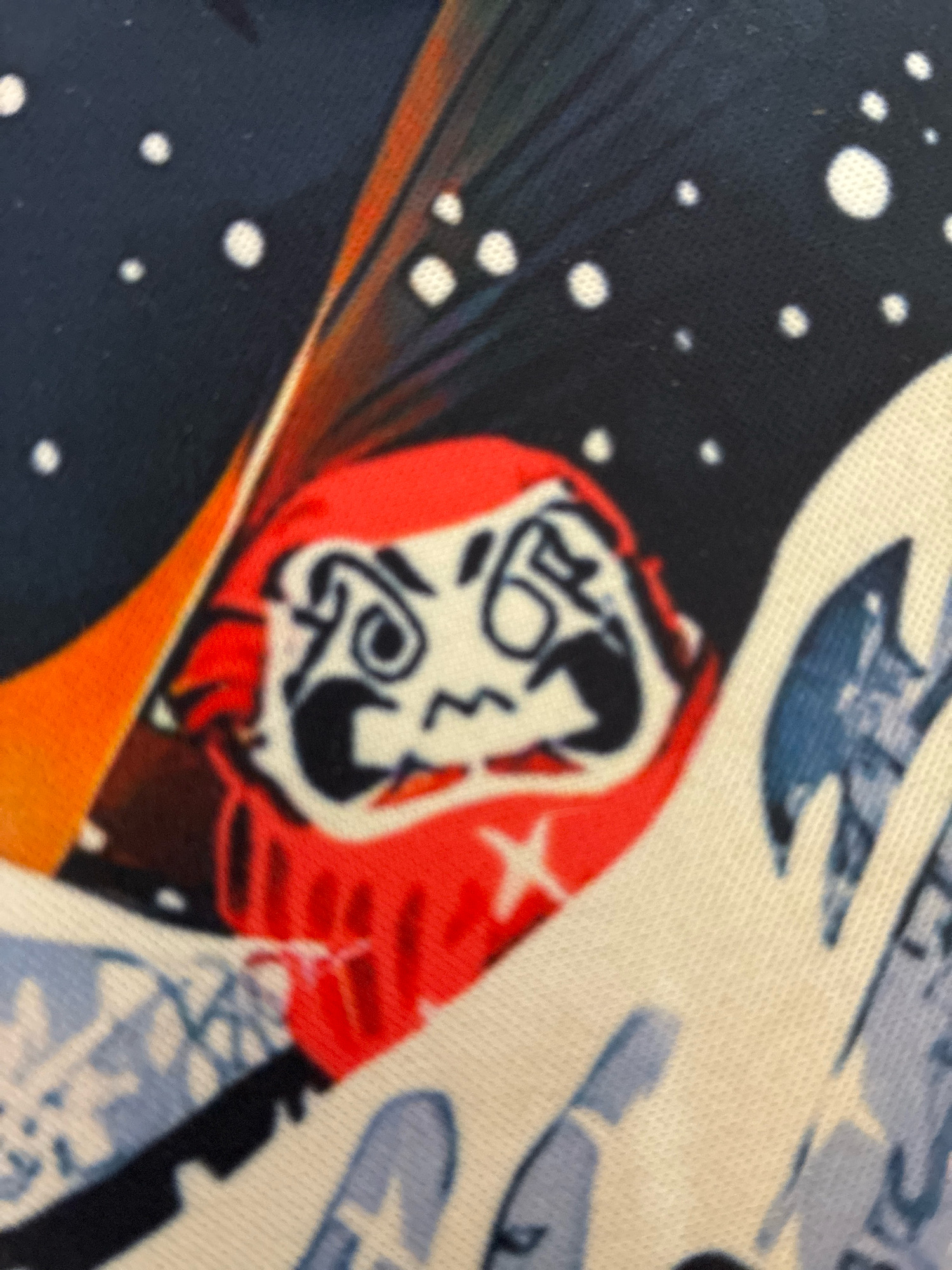 The image is a close-up, slightly blurred shot of a red Daruma doll against a dark blue background with white dots, possibly representing stars. The doll has a white face with black features, including exaggerated eyebrows, eyes, and a mouth. To the left of the doll, there are streaks of orange and brown, and to the right, there are white and blue wavy patterns, possibly representing water or fabric. This image is not from Curitiba, Brazil.