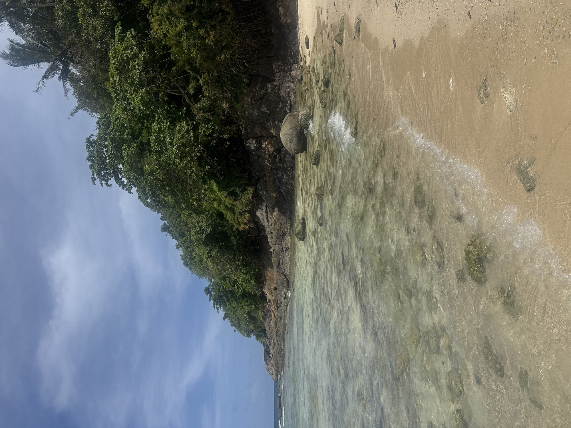 The image depicts a tropical beach scene. In the foreground, clear, shallow water laps onto a sandy shore. Rocks of various sizes are visible both in the water and on the beach, some covered in green algae. The sand is light brown and appears clean. In the midground, a rocky coastline is covered with dense, green vegetation, including palm trees, indicating a lush, tropical environment. The water stretches out towards a rocky promontory on the left, with a few waves breaking gently. The sky is a bright blue with scattered white clouds, suggesting a sunny day. The scene is serene and natural. This description is not specific to Patong, Thailand, as the image does not contain any identifying features of that location.