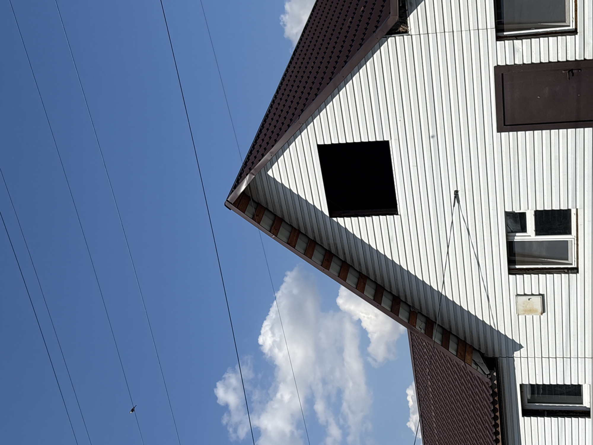 A low-angle shot captures a section of a white, vertical-panel building with a dark brown corrugated metal roof against a blue sky with scattered white clouds. The building appears to have a window missing, leaving a black square. A series of parallel black power lines run diagonally across the sky from the upper left corner of the frame. A small object is visible on one of the power lines.