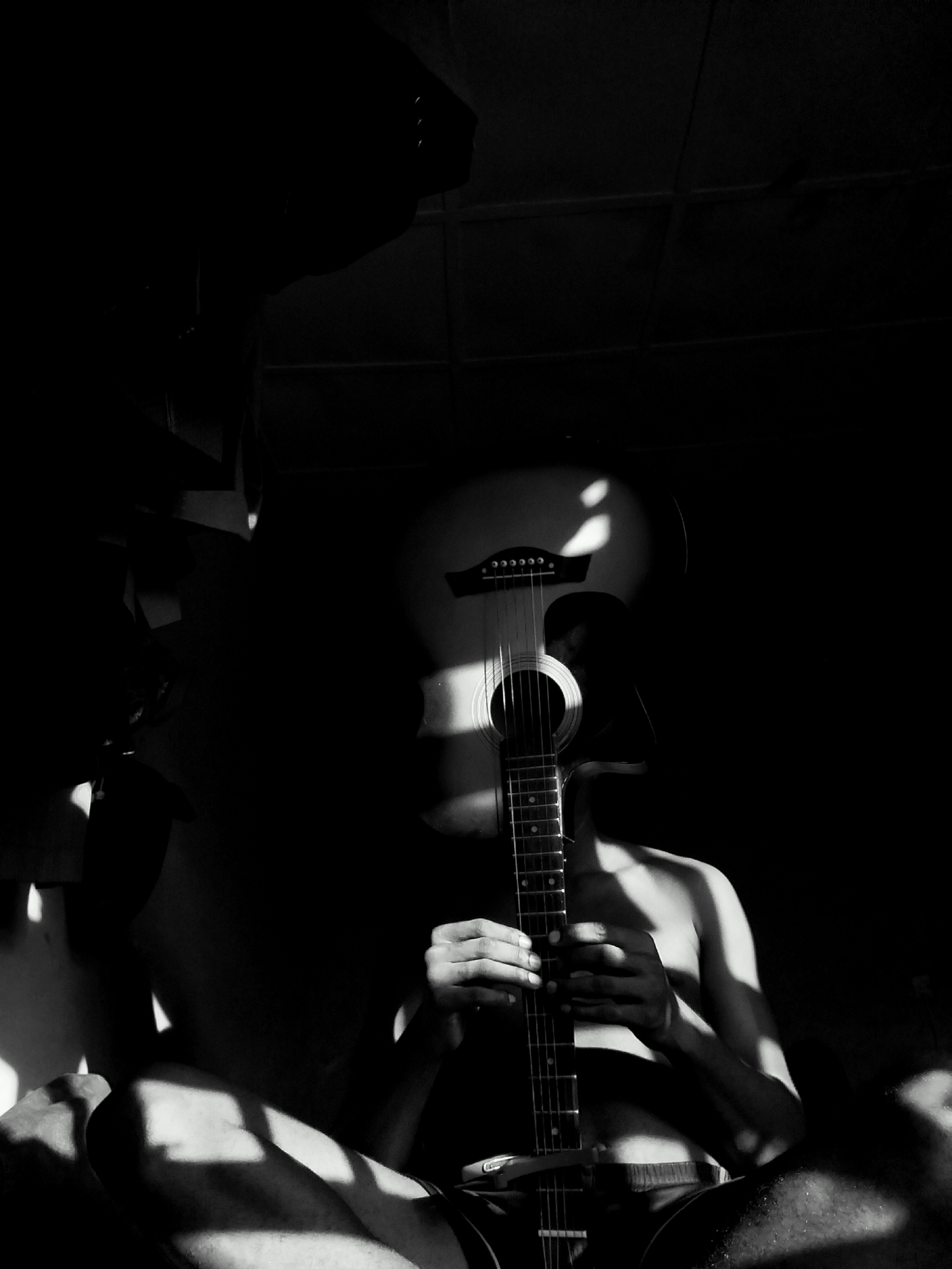 A black and white photograph depicts a person sitting cross-legged, holding an acoustic guitar that obscures their face. The person appears to be shirtless and is wearing shorts. The lighting is dramatic, with stark contrasts between light and shadow. The scene appears to be indoors. There is no information that suggests the image was taken in Keffi, Nigeria.