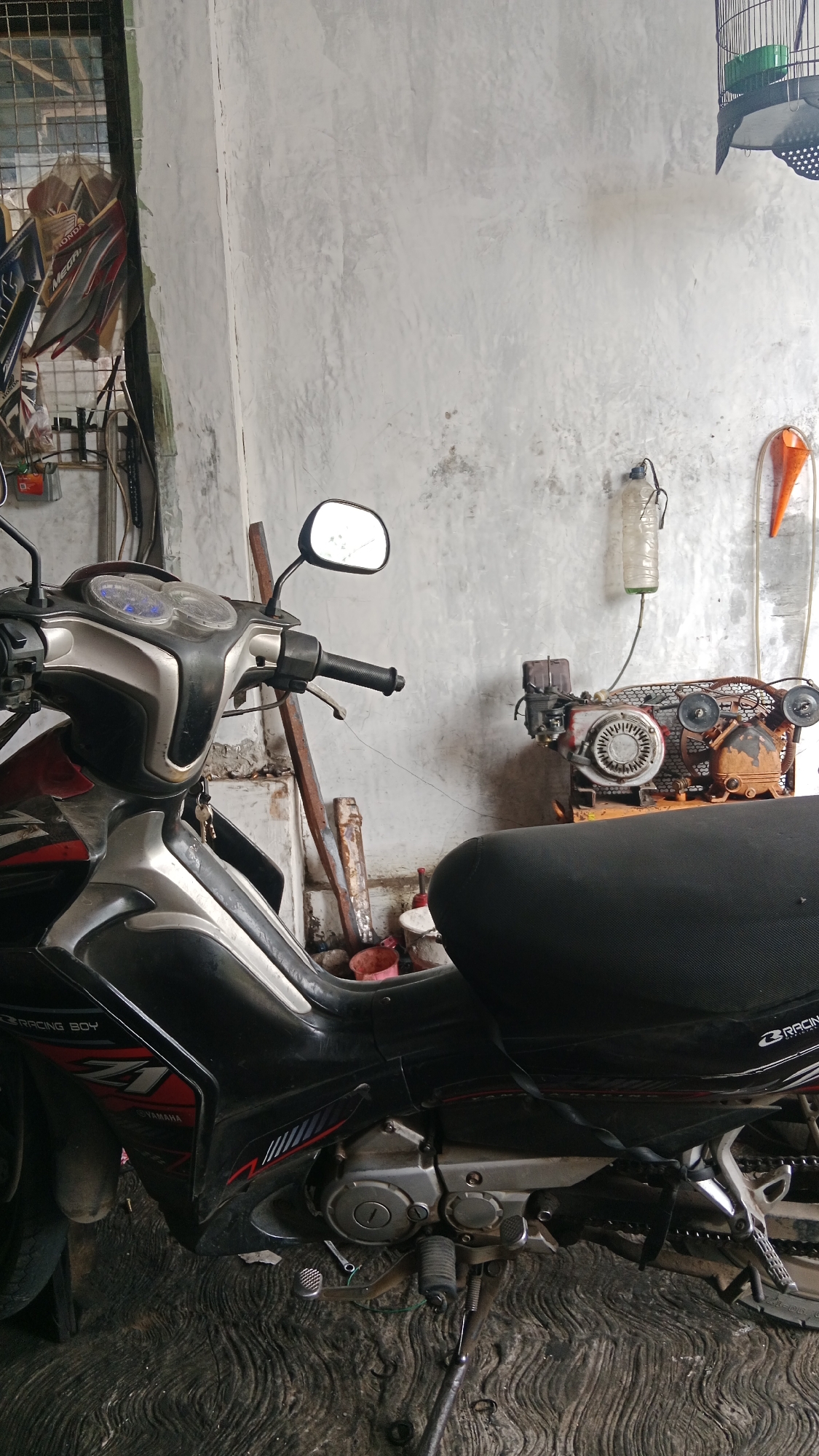A motorcycle is parked in a workshop. The motorcycle is black and silver with red accents and has a black seat. To the right of the motorcycle is an orange cone and a clear plastic bottle hanging from a string. In the background, there is a metal cage hanging from the ceiling, and a compressor with a motor. The walls of the workshop are concrete and the floor is made of rough concrete. The scene appears to be located in the city of Candigugur, Indonesia.