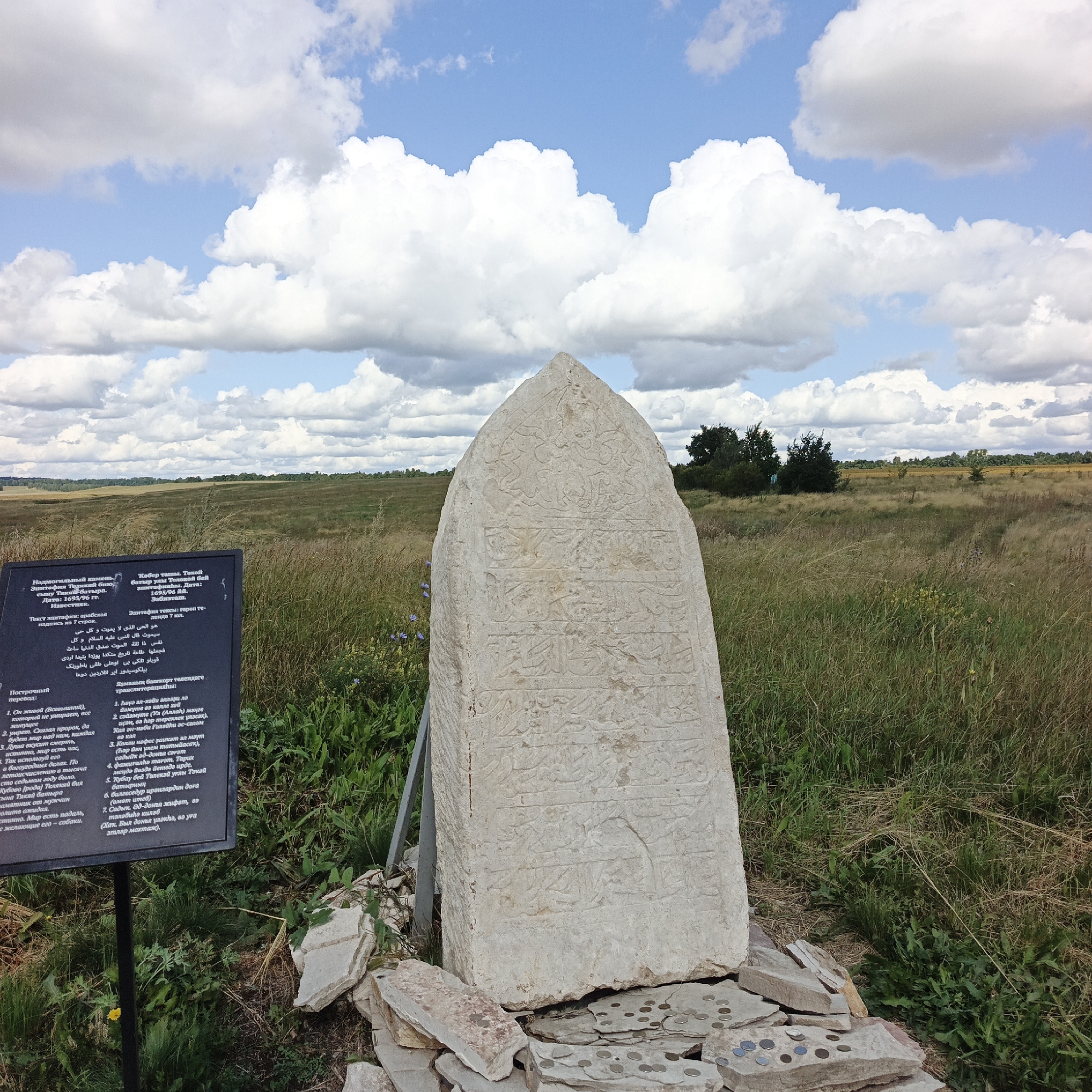 In Verkhnie Termy, Russia, a carved stone obelisk stands in a grassy field under a bright blue sky with scattered white clouds. The obelisk, weathered and with inscriptions, is surrounded by broken stones, some of which have coins placed on them. To the left of the obelisk, a dark informational sign with text in multiple languages is partially visible. The background shows a gently rolling landscape with trees and vegetation. The scene appears to be an outdoor historical site or monument.