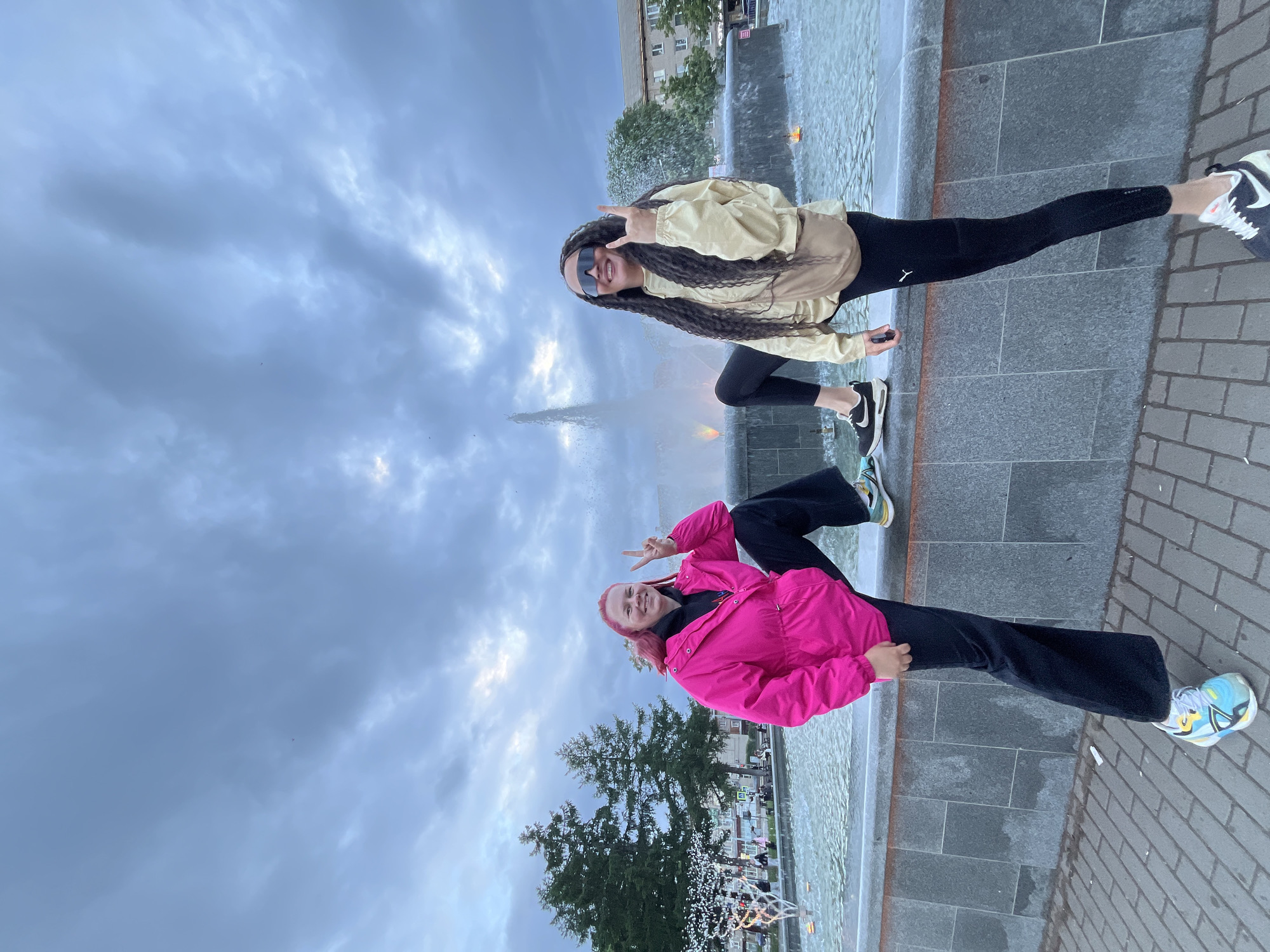 Two young adult females are depicted posing next to a large, multi-tiered fountain in a public square or park in the city of Chaikovsky, Russia. The sky above is heavily overcast with dense, dark gray clouds.
The woman on the left, with reddish-orange hair, is wearing a bright pink hooded anorak over a dark hoodie, dark wide-leg trousers, and multi-colored athletic shoes (blue, yellow, black, white). She is smiling and making a peace sign with her right hand, while her left foot is placed on the gray stone rim of the fountain and her right foot is on the brick-patterned pavement.
The woman on the right, with long, dark, textured hair and dark wrap-around sunglasses, is wearing a light beige windbreaker jacket, black leggings, and dark athletic shoes with white soles. She is smiling and making a "rock on" hand gesture with her right hand, while her right foot is placed on the fountain's gray stone rim and her left foot is on the pavement. A light brown fanny pack or crossbody bag is worn across her chest.
The fountain features a tall central water jet and several smaller streams. The base of the fountain is constructed from dark gray stone, with a lighter gray coping stone forming the rim. Orange and yellow lights are visible within the fountain's water. To the far left in the fountain's basin, a decorative illuminated structure, possibly resembling a stylized tree, is present.
In the background, to the left of the fountain, stand several tall, dark green coniferous trees. Further back, on the right side of the image, multi-story buildings of a light color, consistent with urban architecture, are visible. The ground in the foreground is covered with light-colored, brick-patterned pavement.