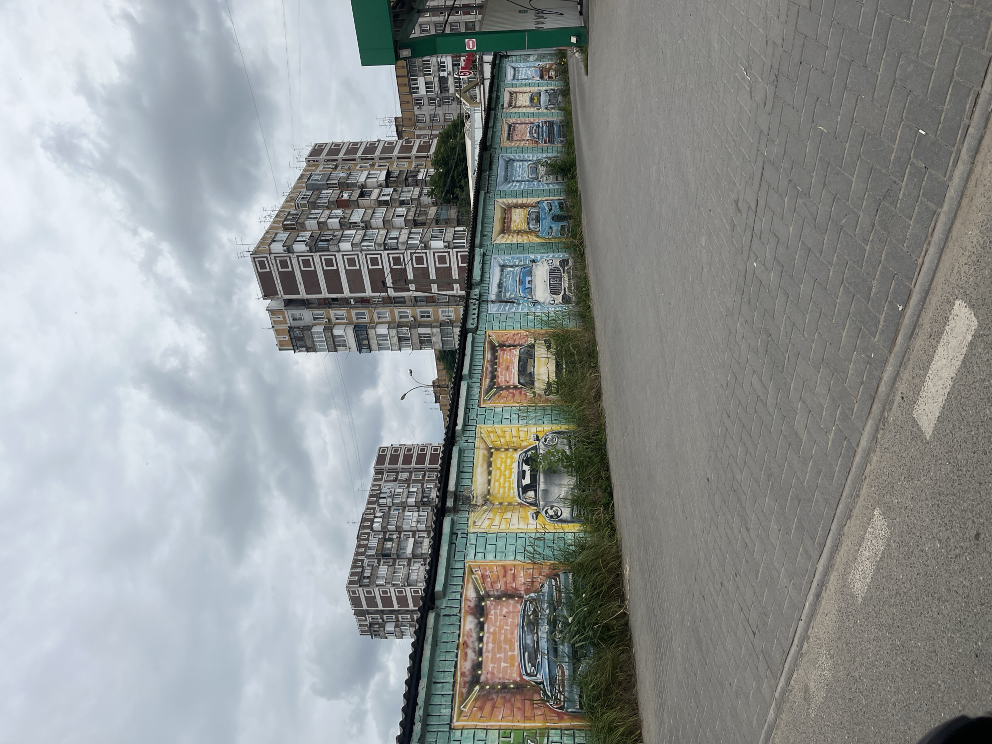 The image depicts a mural of cars painted on a row of garage doors, likely in a residential area. The garages are painted in vibrant colors, with each door featuring a different model of car, as if parked inside. The mural is set against a backdrop of overcast skies and typical Soviet-era apartment buildings, characteristic of many Russian cities. The presence of a gas station canopy on the right suggests the garages are located near a commercial area. The scene is a common sight in Krasnodar, Russia, where such artistic expressions are often used to beautify urban spaces.