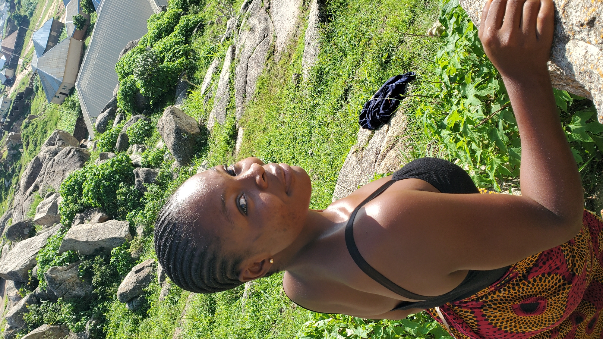 A young Black woman with braided hair is pictured from the shoulders up, looking over her right shoulder towards the camera. She is wearing a black spaghetti strap top and a colorful patterned wrap skirt. She has a subtle smile and appears to be in an outdoor environment with a rocky, grassy terrain and some vegetation. In the background, several buildings with corrugated metal roofs are visible, suggesting a semi-urban or rural settlement. This scene is consistent with the landscape found in the city of Jos, Nigeria, known for its rocky outcrops and elevated terrain. A dark blue piece of clothing lies on the ground behind her.