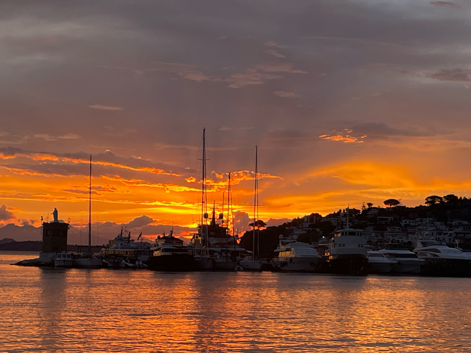 This image depicts a harbor at sunset in Bagni, Italy. The sky is a vibrant mix of orange and purple hues, with scattered clouds. Silhouetted against the sunset are several boats and yachts docked in the water. In the foreground, the water reflects the warm colors of the sky. On the left side of the image, a small lighthouse-like structure with a statue on top is visible, along with a few smaller boats. The background features a hillside town with buildings illuminated by the fading light. There are no people visible in the scene.