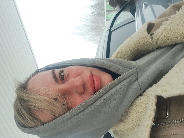 A person wearing a gray hooded sweatshirt and a tan shearling-lined jacket is positioned near a car with a light-colored building visible in the background. The individual's face, slightly red, is prominent within the hood. A tree with bare branches is seen through the car window. The photograph appears to be taken during the day with overcast lighting.