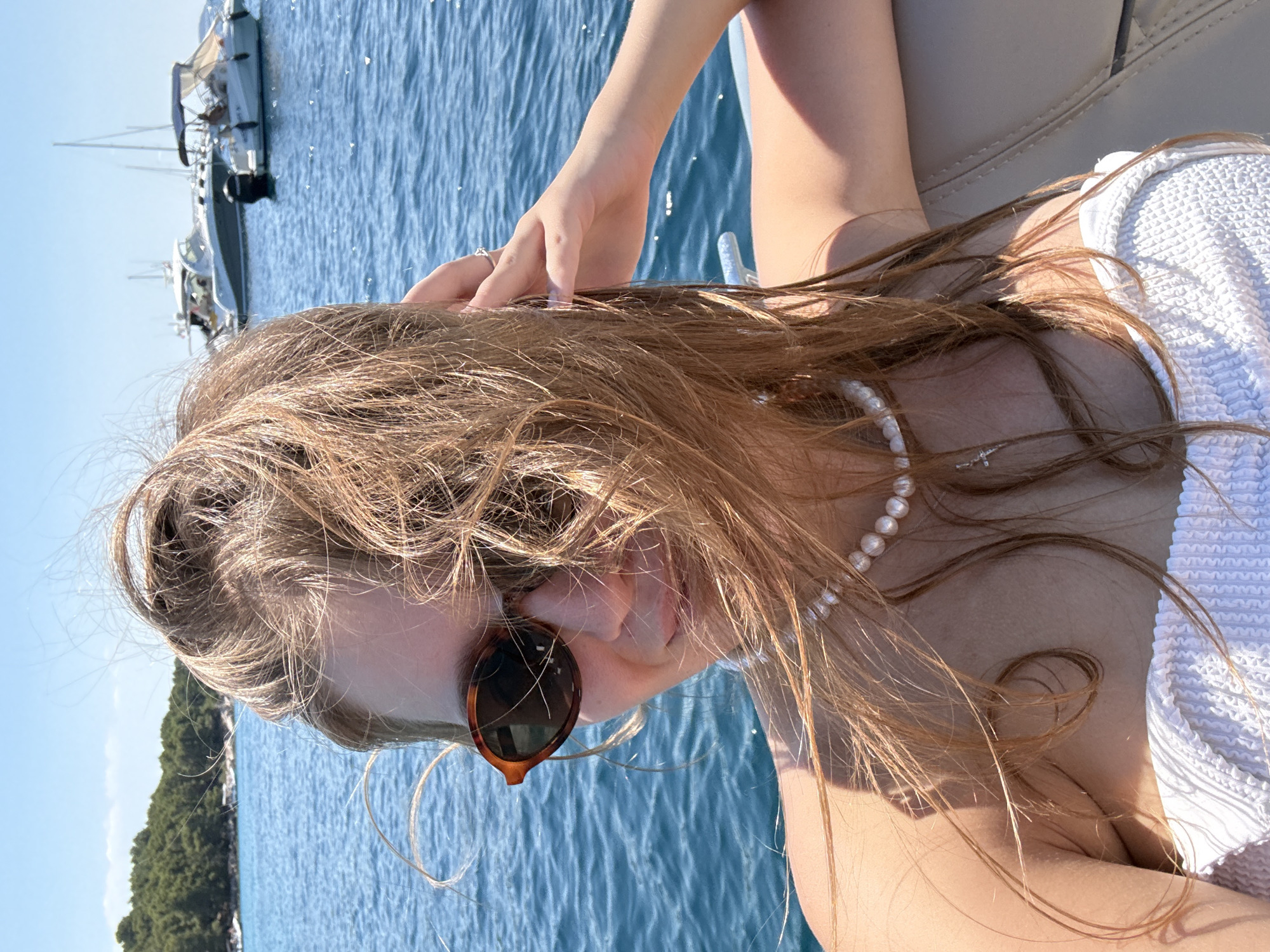 A young woman with long, wavy, blonde hair and sunglasses is taking a selfie from a boat. She is wearing a white bikini top and a pearl necklace. Her hair is blowing in the wind. The water is blue and there are other boats and an island in the background. The scene appears to be a sunny day on the Adriatic Sea, possibly near Vodice, Croatia.