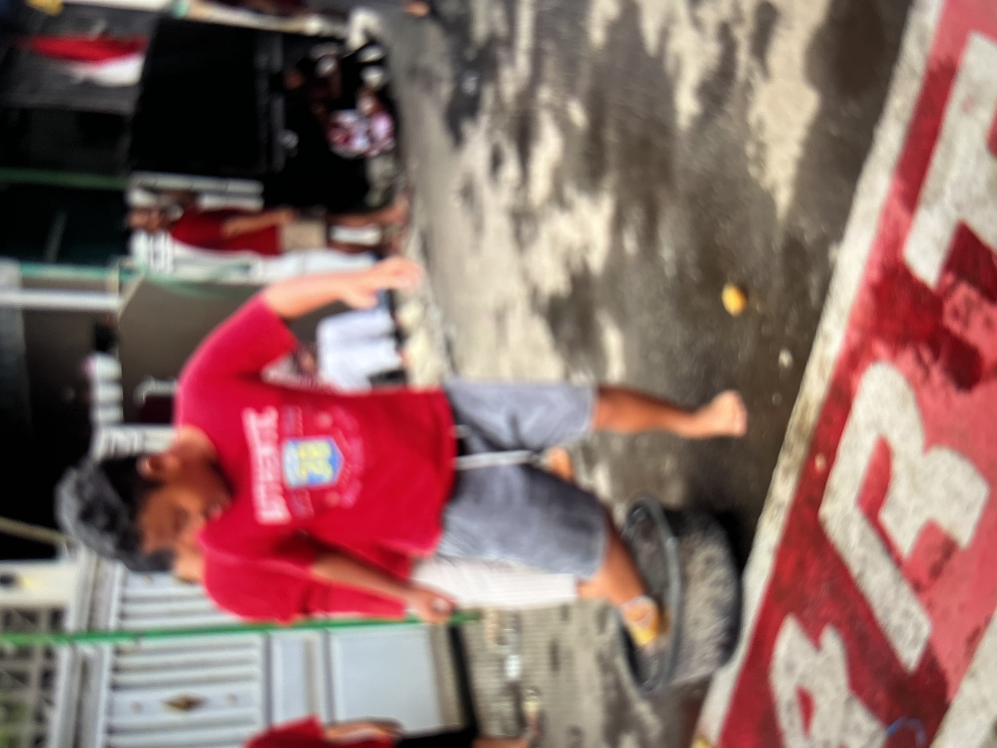 A young boy wearing a red t-shirt with "AUTHENTIC" printed on it and grey shorts is standing with his right foot in a dark-colored bucket filled with water. He is participating in an activity that appears to be a game or competition, as indicated by the red and white striped banner in the foreground with white lettering, possibly spelling out "BARONGSAI" or a similar word, which is common in Indonesian cultural festivals. The boy's left foot is bare, and his body is angled as if he is either stepping into or out of the bucket.
In the background, other people are visible, including a person in a red shirt and beige shorts standing further back, and another individual partially obscured by the boy. The environment suggests an outdoor setting, possibly a street or public area in Cirahayu, Indonesia, with a building and a fence visible on the left. The ground is wet and appears to be asphalt or concrete, with some puddles and debris. A yellow object, possibly a fruit or marker, is on the wet ground near the banner. The overall scene is somewhat blurred, indicating motion or a low-quality photograph.