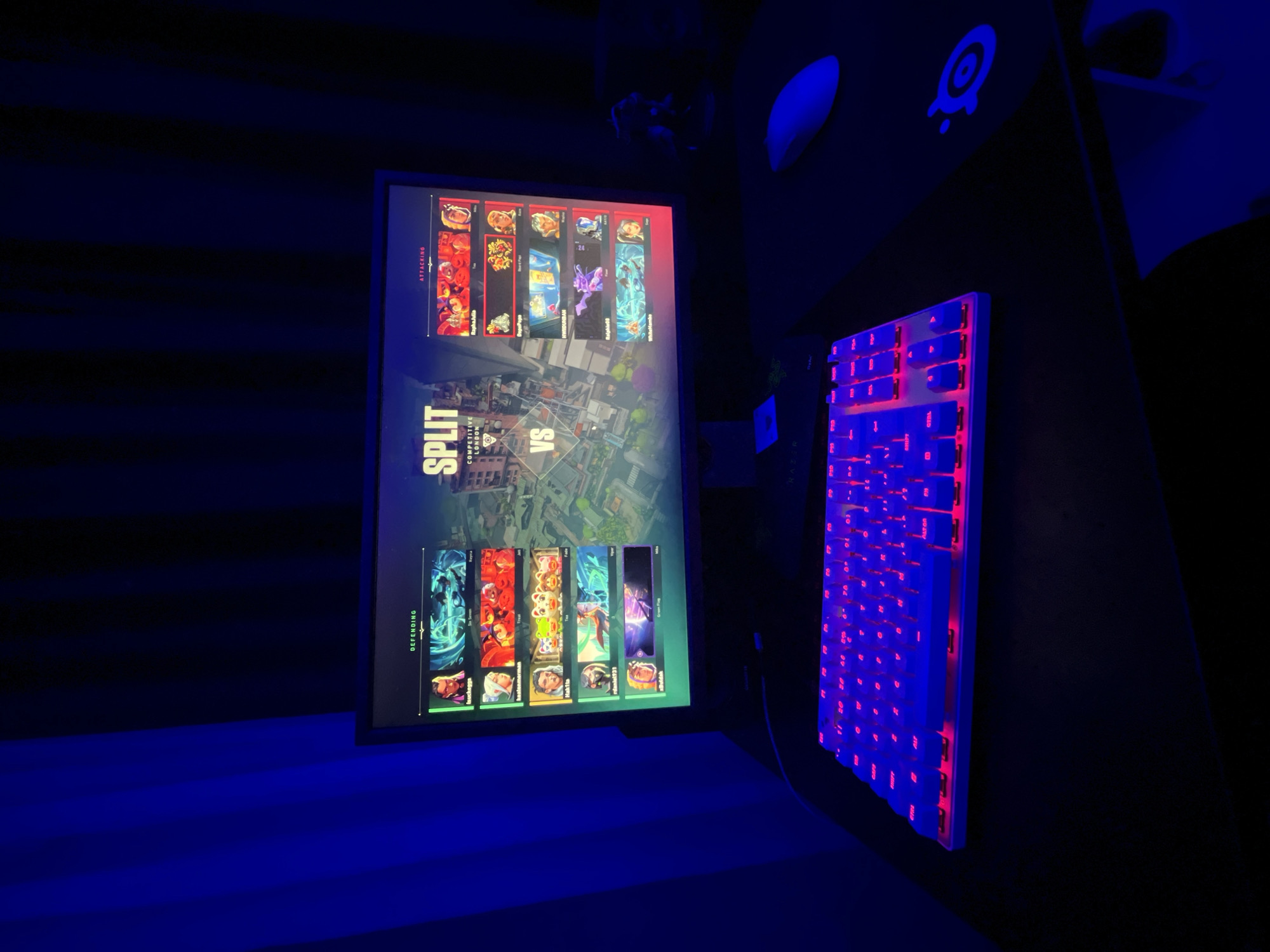 The image shows a gaming setup in a dark room. A monitor displays the agent selection screen from the video game Valorant, showing the map is Split. The teams are Attacking and Defending. The setup also includes a white keyboard with red backlighting. A SteelSeries logo is visible on the desk. No people are visible. There is no evidence to suggest the location is in Burgas, Bulgaria.