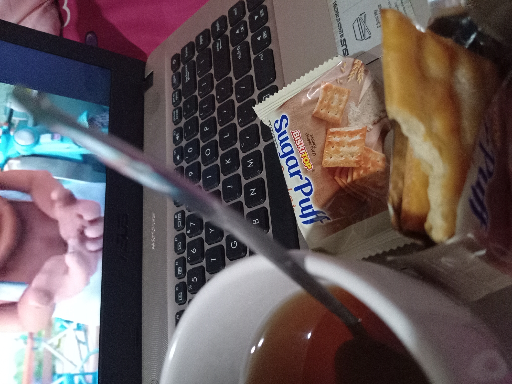 The image shows a close-up view of a laptop keyboard, a white mug of tea, and a package of biscuits. A piece of food, possibly a pastry or bread, is also visible. The laptop screen displays an out-of-focus image of a person's hands. The scene appears to be indoors, possibly a home or office environment. The ASUS logo is visible on the laptop. The biscuit packaging has text that appears to be in Indonesian, including "BisBiskTOP" and "Sugar Puff". The overall impression is one of casual leisure, with someone enjoying a snack and a drink while using a laptop. The location is specified as Claket, Indonesia.