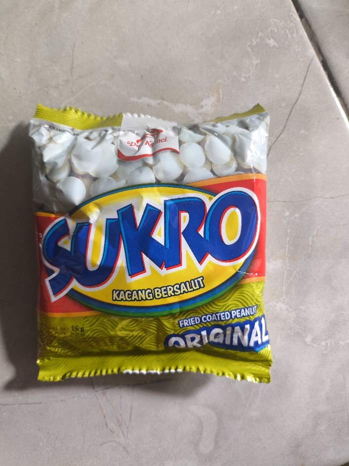 A package of Sukro Original Fried Coated Peanuts sits on a gray surface. The yellow, red, and blue packaging is mostly intact, revealing small, white, coated peanuts at the top. "SUKRO" is prominently displayed in blue with a yellow border, and the text "KACANG BERSALUT" and "FRIED COATED PEANUT ORIGINAL" are visible below. The bottom of the package has a textured yellow pattern.