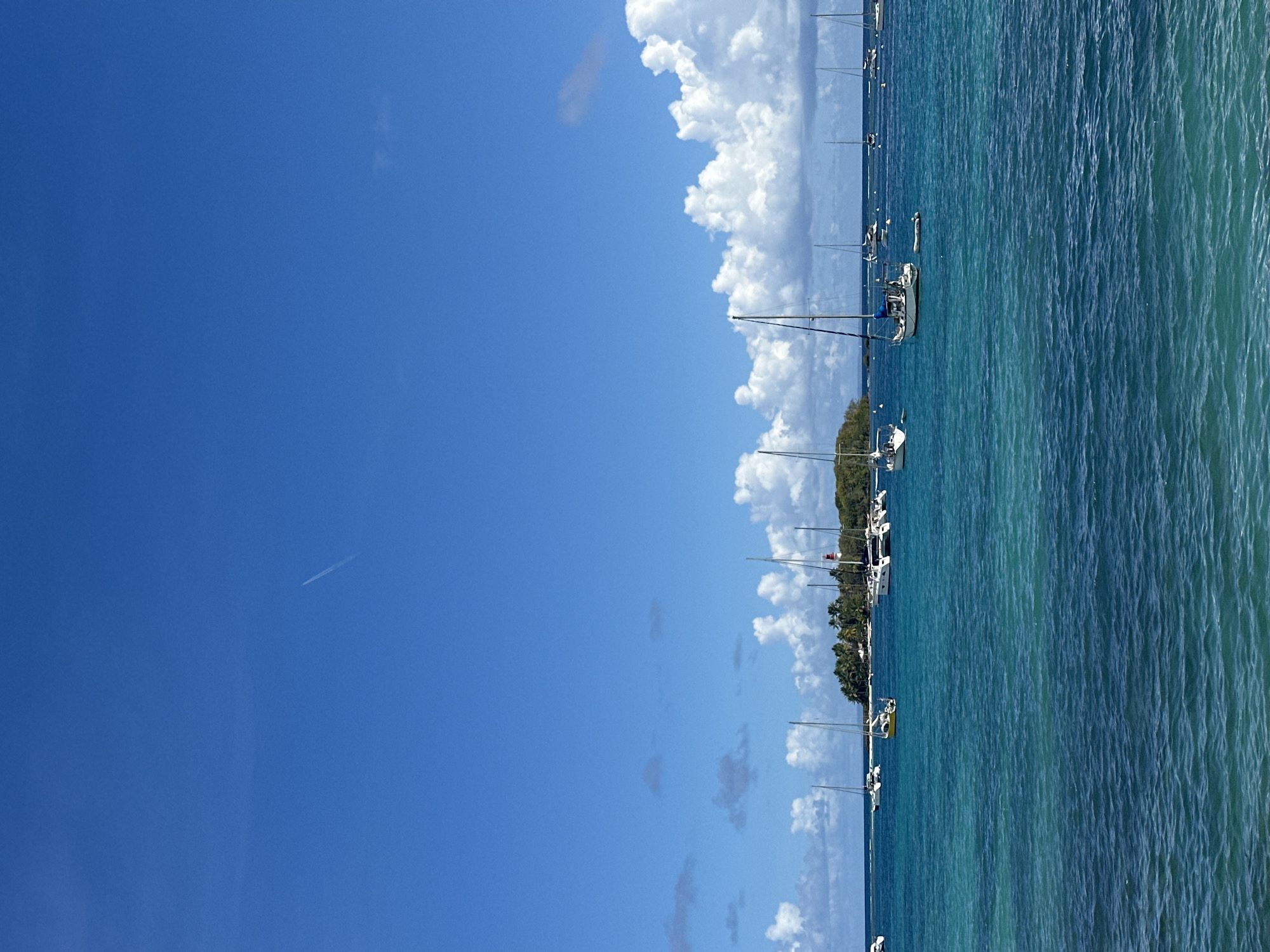 The image shows a scene on the coast of Le Gosier, France. A clear blue sky occupies the left portion of the image, with a faint contrail visible in the upper-left quadrant. Moving toward the right, there's a dense line of cumulus clouds. The right portion of the image features a view of the ocean, with varying shades of blue and green. Several sailboats are anchored offshore, their masts pointing upwards. A strip of land with trees is visible between the line of clouds and the sailboats, likely a part of the coastline. The water is textured, suggesting a light breeze or current.