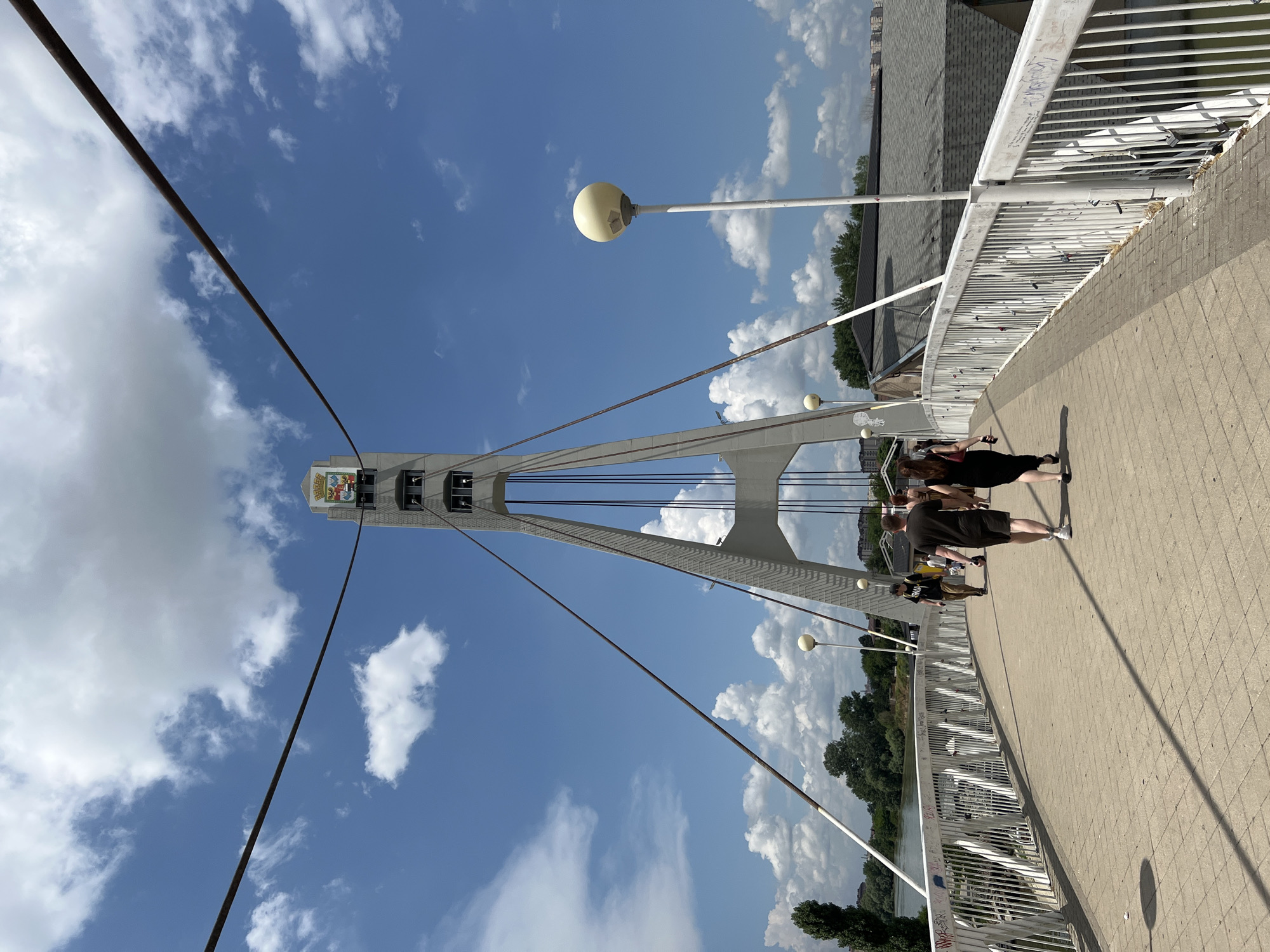 A pedestrian bridge with white railings arches over a body of water. Several people are walking across the bridge. On the left, a river or canal is visible, lined with trees and buildings. In the background, a tall, concrete tower with cables supporting the bridge rises against a blue sky with scattered white clouds. The tower is topped with a decorative emblem and what appears to be bells. White lampposts with spherical tops line the bridge. The pavement of the bridge is made of interlocking bricks, and shadows from the people and railing are cast across it. The railings are adorned with numerous locks.