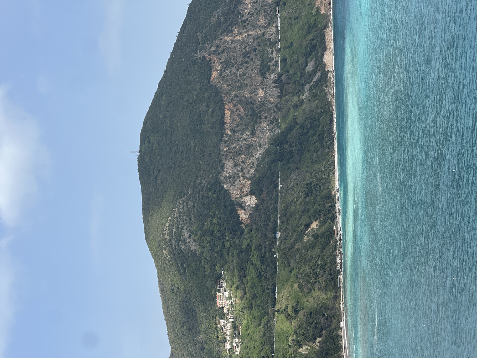 The image depicts a coastal scene in or near Visnjeva, Montenegro. A verdant, steep mountain rises alongside the turquoise-colored sea. A road runs along the base of the mountain, following the coastline. Buildings are nestled on the mountain's lower slopes. A tall communications tower is visible on the mountain's peak. The sky is clear blue.