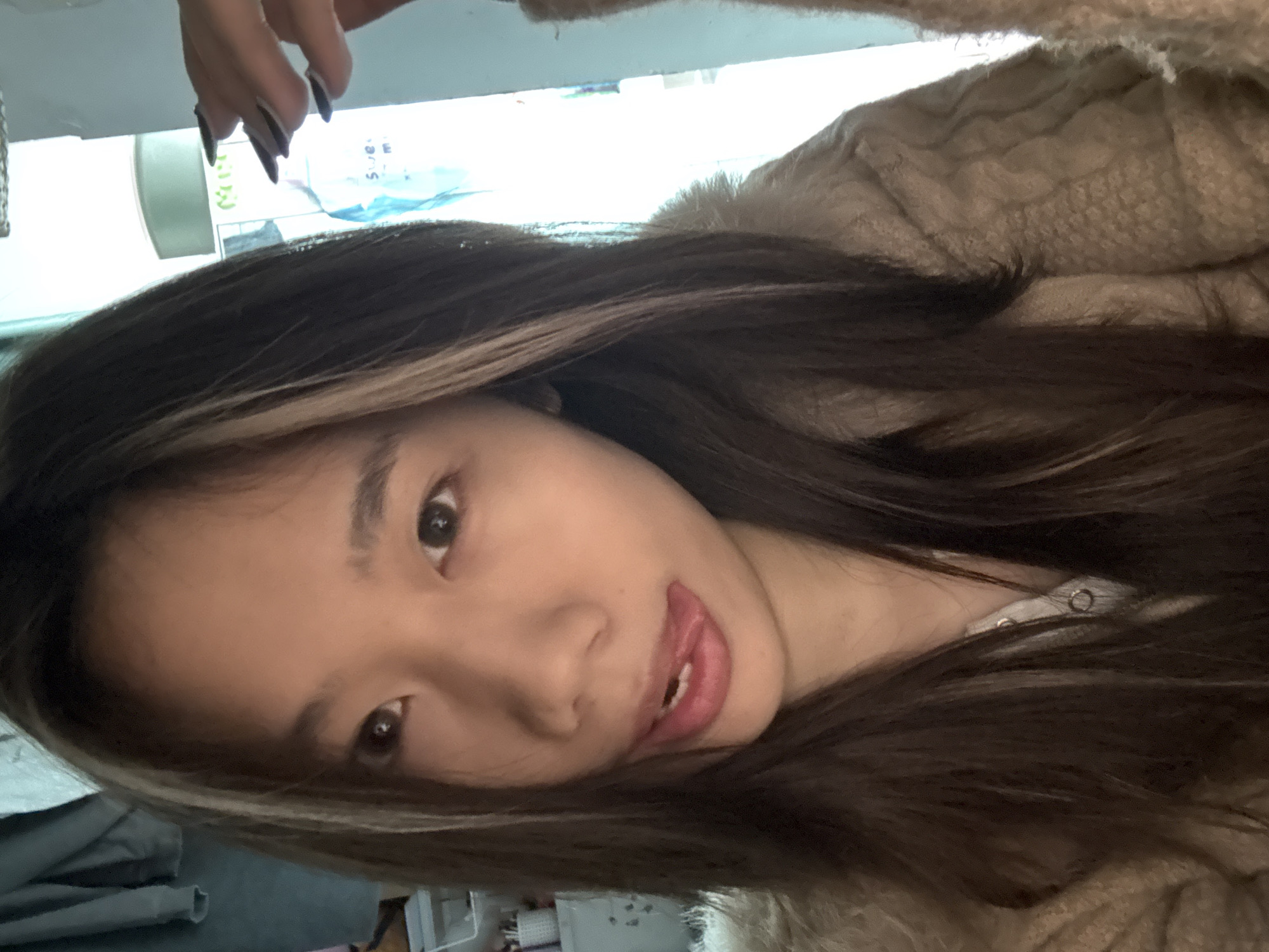A close-up indoor photograph depicts a single East Asian female subject with long, dark hair featuring prominent lighter streaks near the face. She is looking directly at the camera with her tongue slightly extended from her mouth, revealing the tip and lower incisors. She wears a light-colored, possibly beige or tan, textured outer garment, appearing to be a knitted or fluffy cardigan or jacket, over a lighter-toned undergarment with visible snap buttons at the neckline. In the upper right quadrant, a hand with long, dark, pointed fingernails is partially visible. The background on the left side reveals a cluttered area with shelving holding various small items and a segment of a light-colored, possibly furry or soft, object. The right side of the background is brightly illuminated, showing indistinct light-colored objects and reflections, including text partially readable as "Swe..." and "m". No specific visual cues within the image allow for the determination of a precise location within Nan'an District, China; the scene appears to be an interior domestic space.