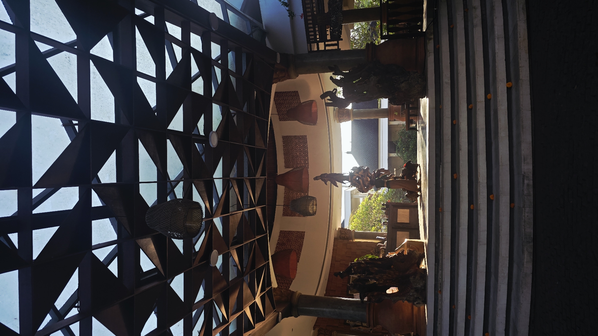 The interior of a building in Ubud District, Indonesia, features an elaborate ceiling with a geometric pattern of dark triangular shapes and open spaces revealing the sky. Wicker lanterns hang from the ceiling. Decorative elements include woven wall hangings and wooden sculptures. Stone pillars support the architecture. A staircase with orange flowers on each step leads to an outdoor area with trees and a building visible in the background.
