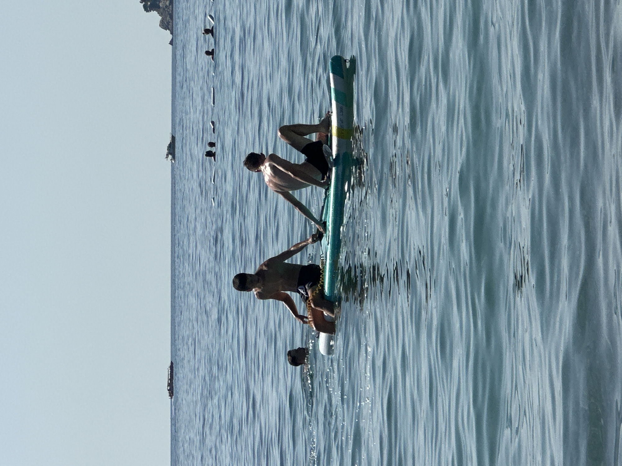 In the waters near Đurovići, Montenegro, two young males are engaged in water activities. One is sitting on a paddleboard, his legs bent and arms resting on his knees. The other, partially submerged, is swimming nearby. In the background, several other individuals are also in the water, some with paddleboards. Further out, two boats are visible on the horizon, under a clear sky. The sea is calm with gentle ripples.
