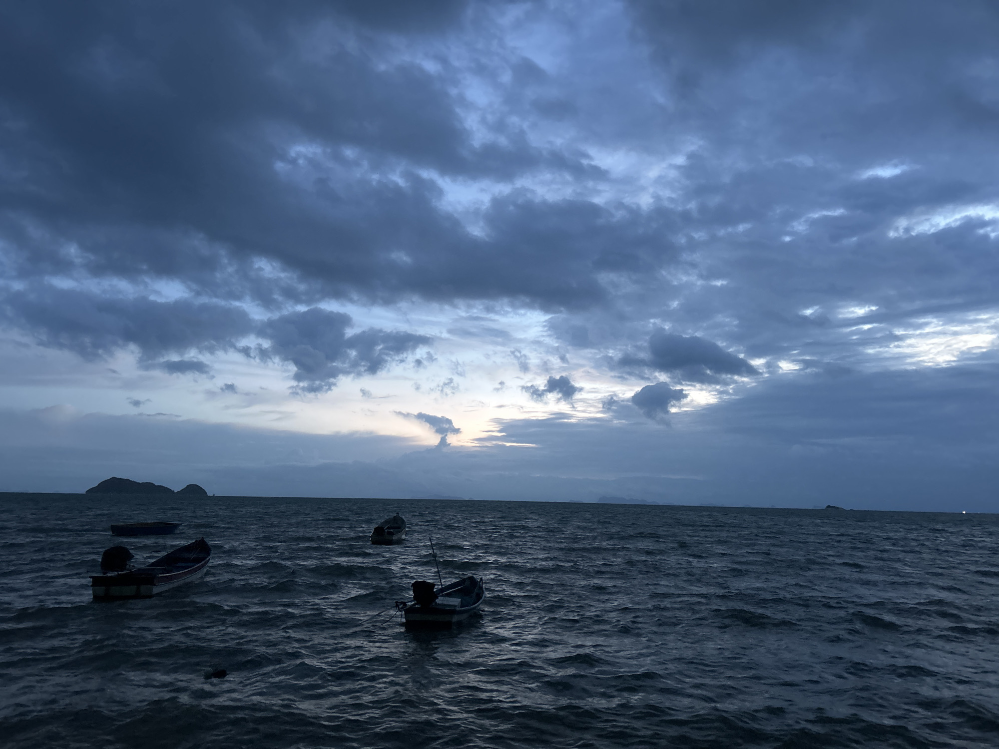 The image depicts a body of water, likely the sea, with several small boats adrift. The sky is overcast with dark, dramatic clouds, suggesting either a storm is approaching or has recently passed. The light is dim, indicating it is likely dusk or dawn. In the distance, islands or land masses are visible on the horizon. The water is choppy, with visible waves. The boats appear to be traditional fishing vessels, some with outboard motors. The overall scene is moody and atmospheric, with a sense of quiet solitude. The location is described as Ko Pha-ngan District, Thailand.
