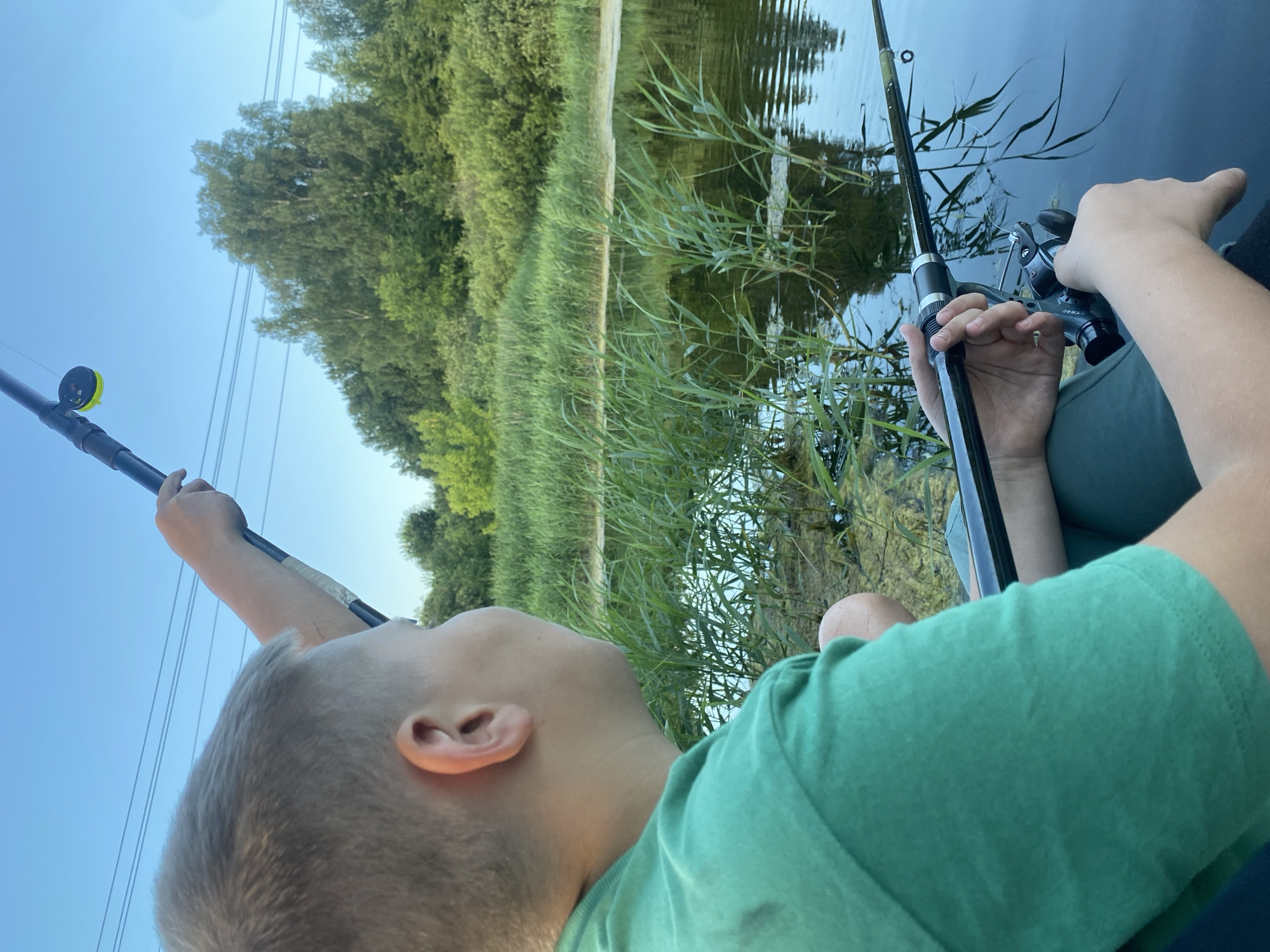 A boy, seen from the back and right side, is holding two fishing rods by the water. He is wearing a green t-shirt and dark pants. The boy is in the foreground and his face is turned to the right, looking towards the water. The water is calm and reflects the sky and trees. There are reeds growing in the water. In the background, there are trees and a clear blue sky. The scene appears to be set in a natural environment, possibly a lake or river, and given the context of the city of Afanasyevo, Russia, it is a rural setting outside of the city. The lighting suggests it is either early morning or late afternoon. There is no indication of any other people or animals in the image.