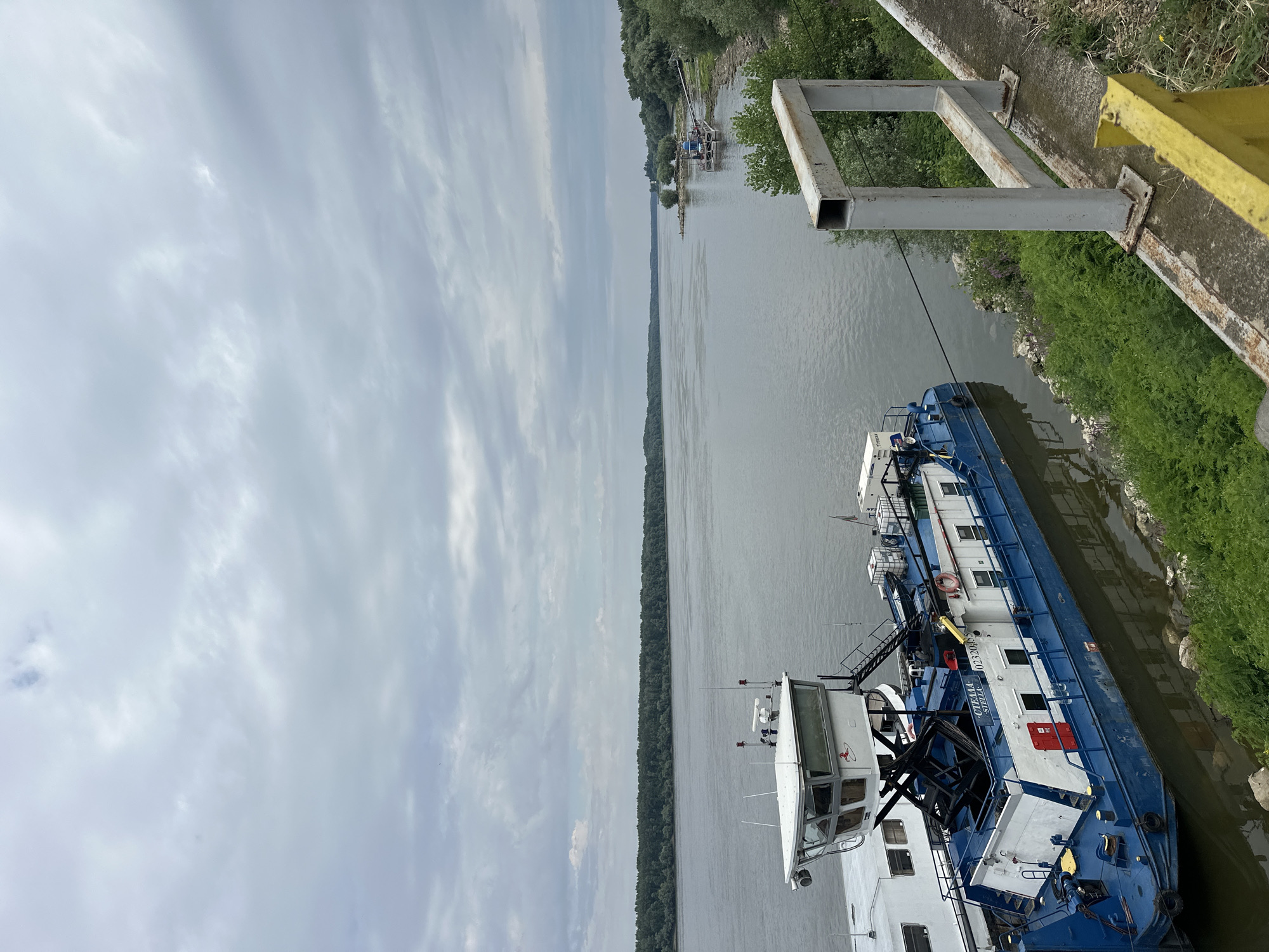 The image depicts a cloudy day along a wide river, likely the Danube, near Marten, Bulgaria. In the foreground, two barges are docked. The larger barge is blue and white, with "STELLA" visible on its side and the number "02320455". It has a prominent white cabin structure elevated on a metal frame. The second barge, partially visible, is mostly white. The river is calm and stretches towards a distant, tree-lined shore. On the right side of the image, a concrete embankment runs along the riverbank, with green vegetation and a metal railing. There are no people visible in the image.
