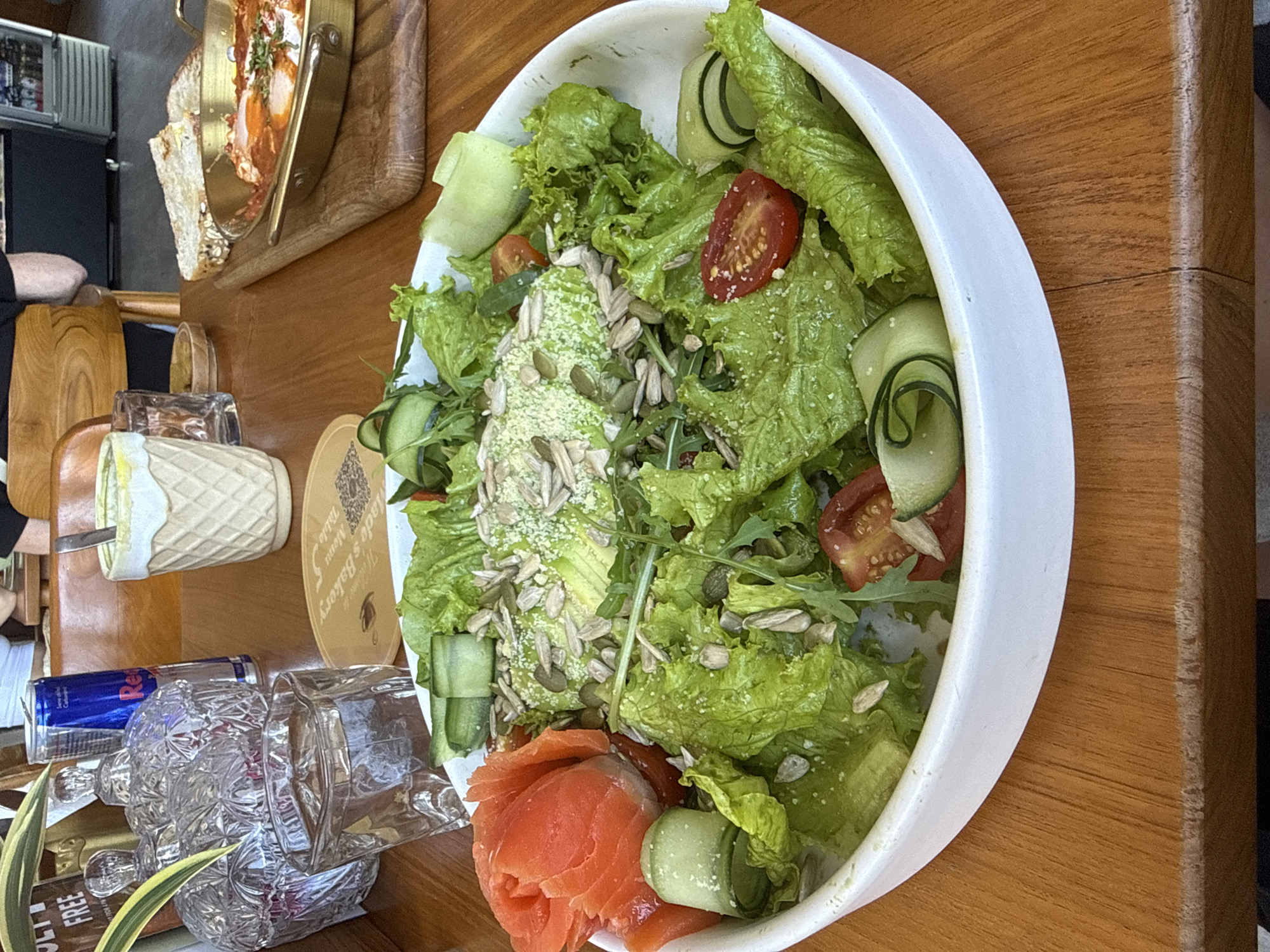 The image shows a table at a restaurant in Ungasan, Indonesia. A white bowl filled with a salad dominates the right side of the image, containing lettuce, sliced cucumbers, tomato wedges, smoked salmon, and seeds. Adjacent to the salad bowl, on the left, are a glass of water, a Red Bull can, and a small ceramic container with a spoon, possibly holding dressing or sauce. In the upper-left corner, a wooden serving board holds a dish with bread and some kind of egg preparation. The table itself is made of wood.