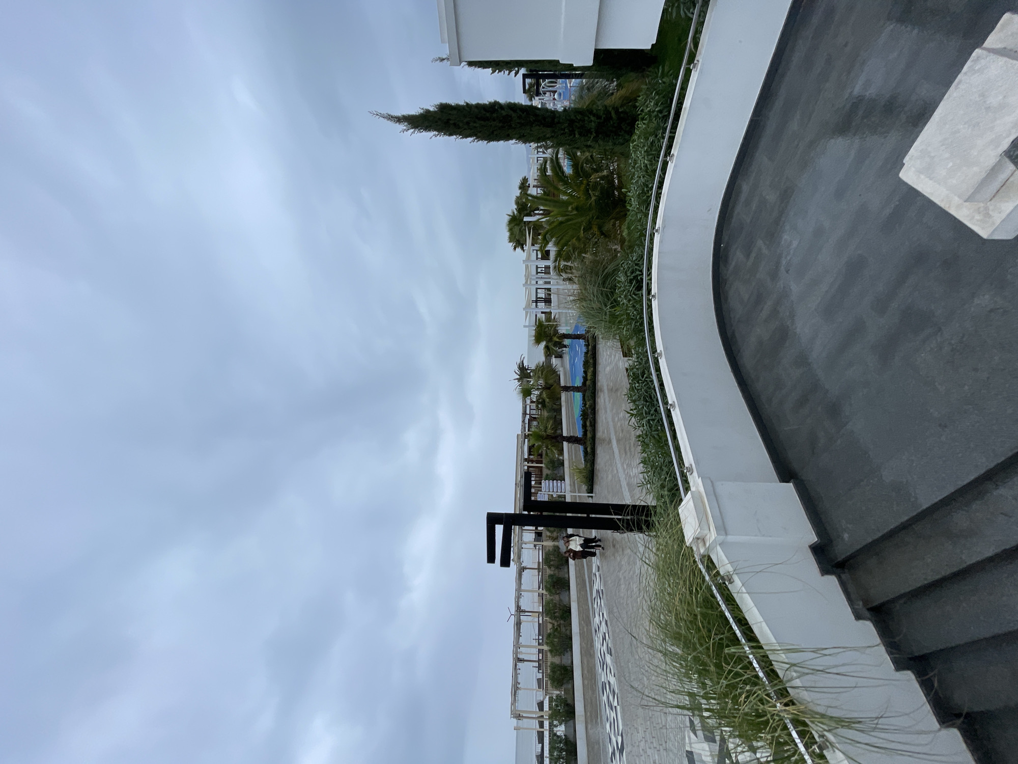 This image depicts a view of a resort or hotel area in Sochi, Russia. The sky is overcast with gray clouds. The architecture features modern white buildings with palm trees and other vegetation interspersed. In the mid-ground, there's a walkway with black and white geometric patterns. A black metal structure stands along the walkway. Two people are present near the structure, but they are small and details are difficult to discern. A curved white wall with vegetation on the side, likely a terrace or balcony, occupies the foreground. The wall has steps leading to a paved area.