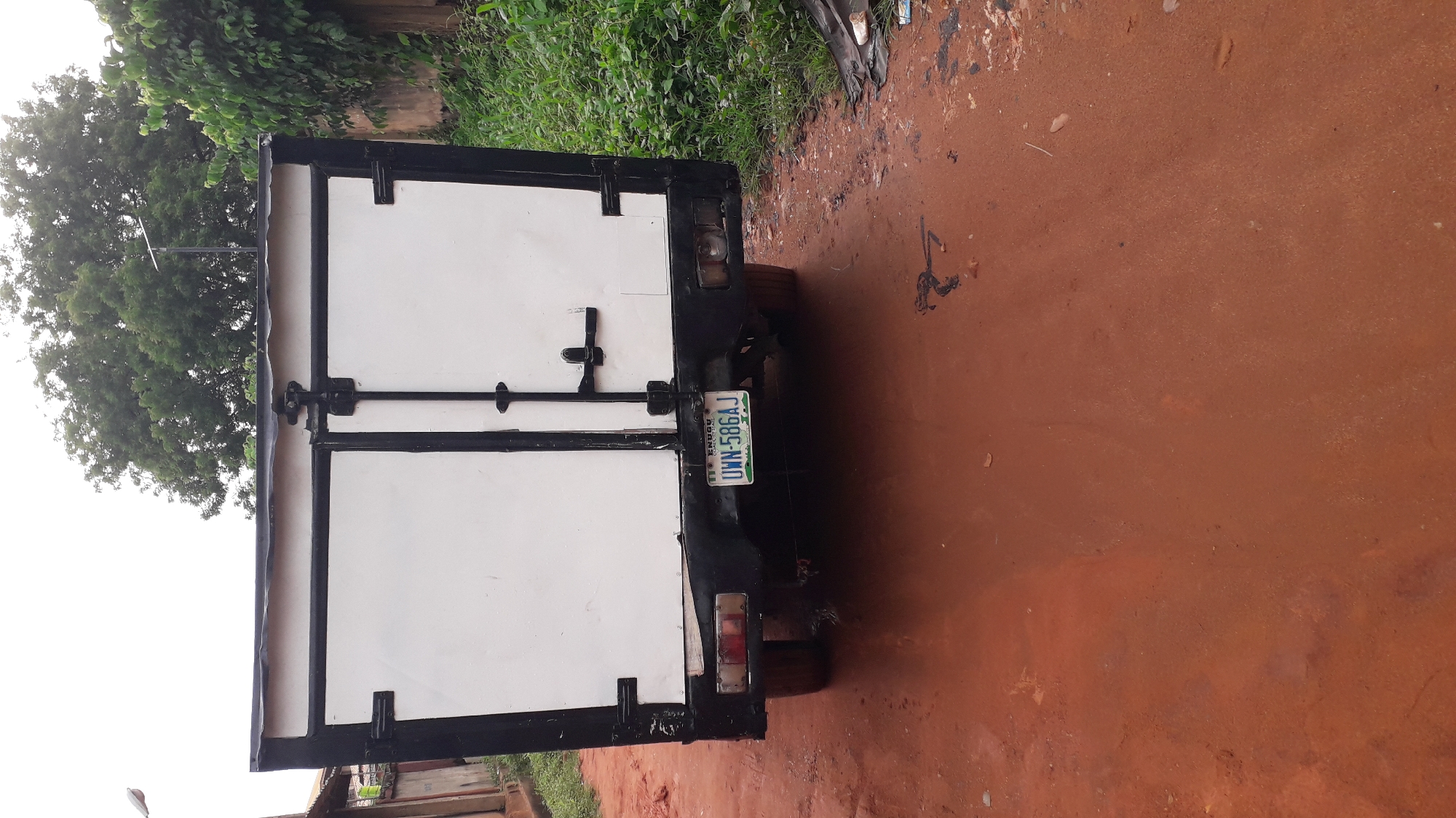 The image shows the rear of a small truck with white double doors framed in black, parked on a red dirt road. The license plate reads "UWN-586AJ" with the "Enugu" state designation of Nigeria visible. The truck appears to be in a somewhat overgrown area with vegetation visible in the background. The location, Onitsha, Nigeria, is only given as information to the bot. No people are visible.