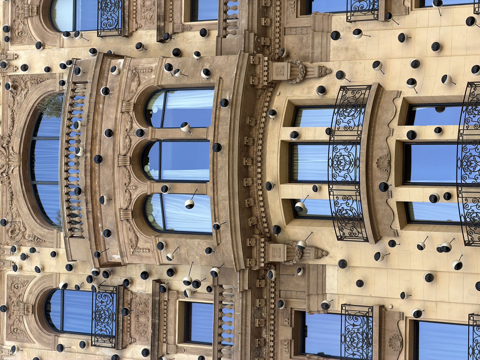 The image showcases the intricate architectural details of a building in Barcelona, Spain, oriented vertically. The facade is adorned with numerous small, round light fixtures, some black and some white with black interiors, attached to thin metal rods. These lights are strategically placed across the building's surface.
The building features several large, arched windows with ornate iron balconies, and other rectangular windows. The windows reflect a clear blue sky, suggesting it's daytime with fair weather. The architectural style appears to be a blend of classical and modern elements, with decorative moldings and textured stonework.
The image captures the building from a close-up perspective, emphasizing the textures and repetition of the decorative elements. The orientation of the image, with the building's facade running vertically, creates an unusual and artistic view of the architecture. There is no visible text in the image.