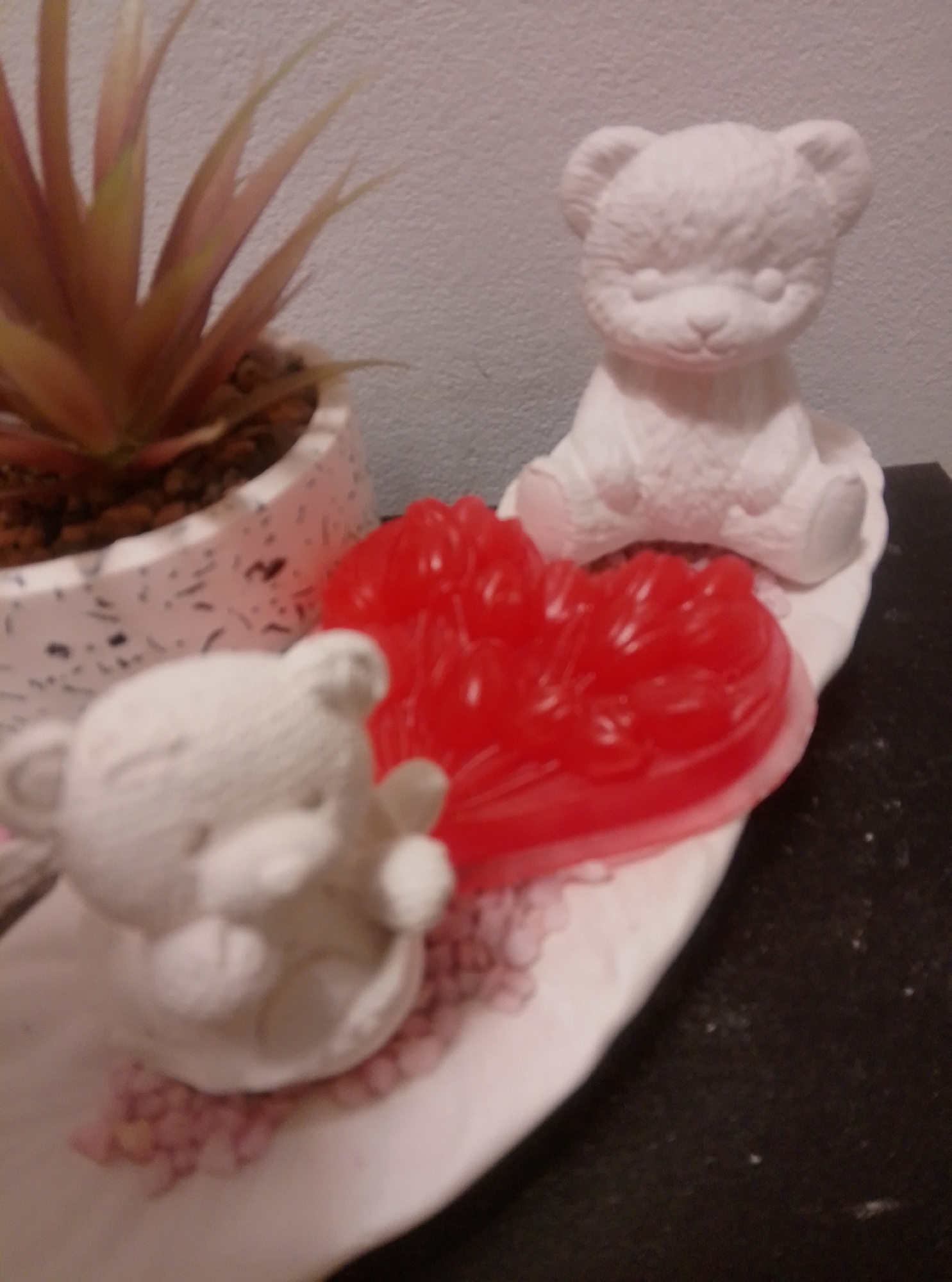 The image shows a still life arrangement on a dark, speckled surface. In the foreground, on the left, is a small, unpainted ceramic teddy bear figure. It is positioned on a white, oval-shaped dish, which is filled with small pink beads. To the right of the teddy bear, on the same dish, is a bright red, heart-shaped object, which appears to be a jelly or candy. Behind the red heart, further back on the dish, is a larger, unpainted ceramic teddy bear figure, sitting upright. To the left and slightly behind the dish is a white pot with a speckled pattern, containing a plant with spiky, reddish-green leaves. The background is a plain, textured light gray wall. The scene is lit from the front, casting soft shadows. The overall impression is of a decorative display.
The location is identified as the city of Wedel, Germany.