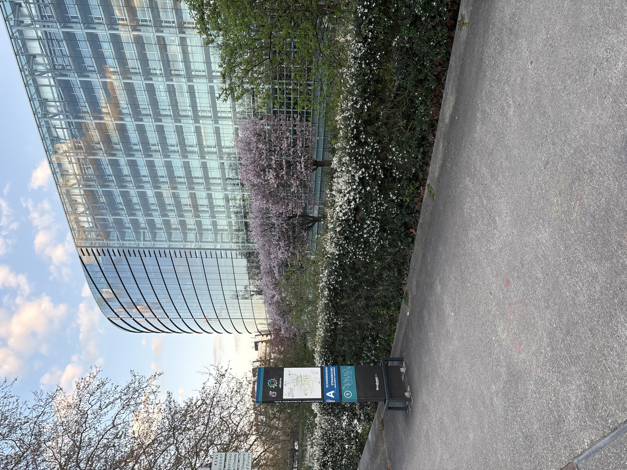 The image depicts a modern glass building, likely an office or government building, possibly the European Parliament in Strasbourg, France, given the visible signage. In the foreground, a public information kiosk with a map and the words "Parlement Europeen Sud," suggesting the European Parliament area. Nearby signage indicates bicycle routes ("Vélos") in the Eckwersheim - Strasbourg region. The kiosk stands adjacent to a concrete sidewalk. There are trees and flowering shrubs in the landscape, including cherry trees in bloom. The sky is partly cloudy.