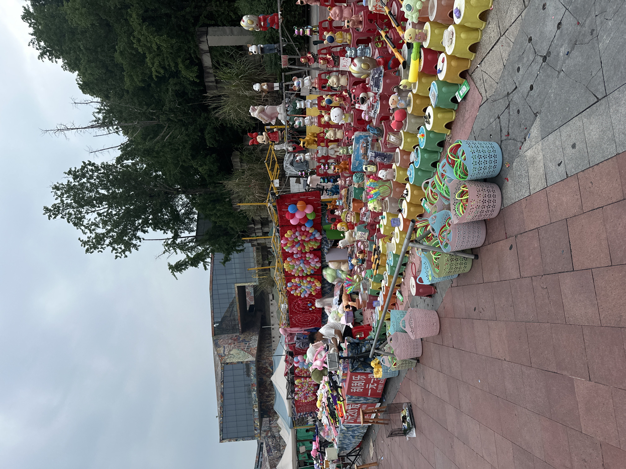 The image shows a street scene, possibly a fair or market, in the Chongqing High-tech Zone, China. A vendor's stall dominates the right side of the frame, filled with colorful prizes and toys. The stall is stacked high with plush toys, plastic containers, and baskets containing colorful loops. A red sign with Chinese characters is visible on the lower right, indicating the items for sale.
In the background, a building with a mural or artwork is partially visible. Trees line the scene behind the building. The sky is overcast. The ground is paved with reddish-brown stones. A person dressed in white is seen at the stall.