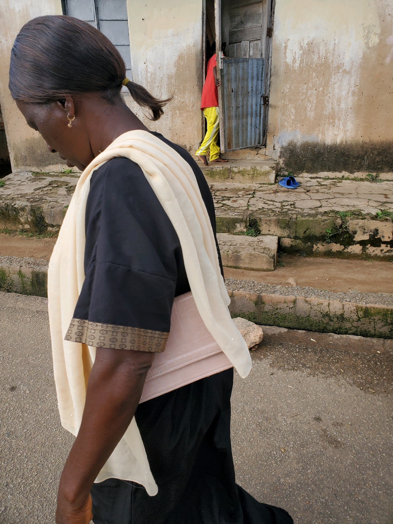 In Jos, Nigeria, a woman with dark skin and her hair pulled into a ponytail walks away from the camera, her head bowed. She is wearing a black garment with a light-colored scarf draped over her shoulders. A patterned trim adorns the end of her sleeves. She carries a light pink rectangular bag tucked under her arm. In the background, a doorway is partially open, revealing a person in a red shirt and yellow pants standing just inside. Blue sandals lie on the ground near the steps leading up to the doorway. The environment appears to be a weathered urban setting with rough concrete steps and textured walls.