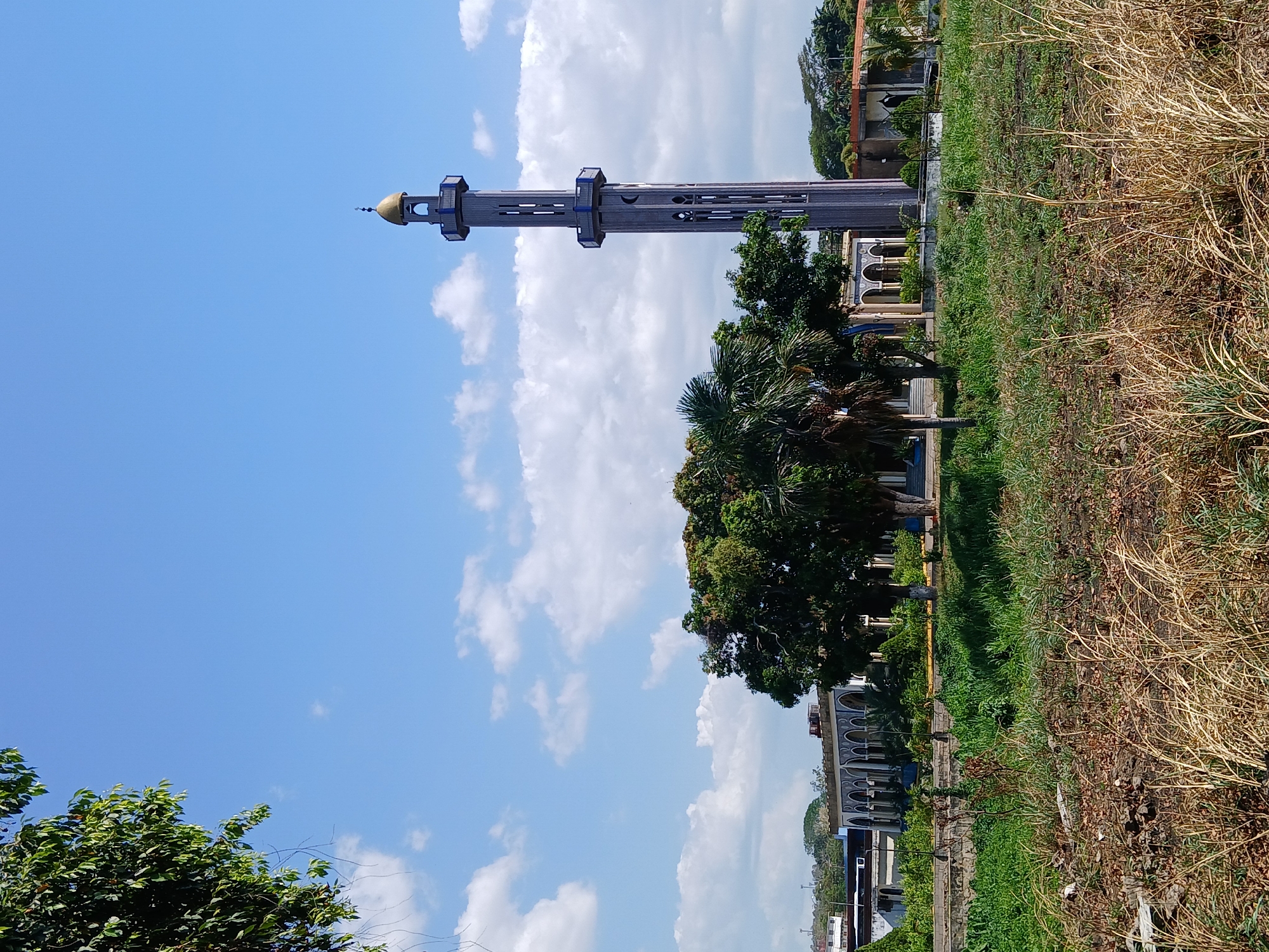 The image shows a minaret of a mosque in Ciudad Guayana, Venezuela. The minaret is a tall, slender tower with a golden dome at the top, built out of gray concrete, and decorated with horizontal blue bands. It is surrounded by green trees and vegetation. The sky is blue with scattered white clouds. The mosque itself has an ornate arched facade and is visible behind the trees. A dry, grassy area is in the foreground, bordering a green lawn closer to the mosque. The overall setting is a sunny day in an urban environment with a focus on the architectural detail of the mosque and its surroundings.