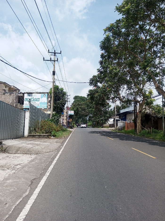 The image displays a straight asphalt road in the city of Khajuripada, India, under a partly cloudy sky. The road features a solid white line on its left edge, delineating it from a cracked concrete sidewalk, and broken yellow lines indicating two traffic lanes further in the distance.
On the left side, a corrugated metal fence runs parallel to the road. Behind this fence, a two-story building with a light-colored facade is visible. A large billboard is mounted on this building, featuring the prominent Indonesian text "ANANTA UMROH" and "23 Jutaan," alongside an image of a man in a white shirt. A wooden utility pole, heavily strung with multiple overhead power and communication lines, stands centrally on the left. Scattered green foliage and small trees are present along this side.
On the right side of the road, a dense line of large trees with green leaves, some bearing reddish-orange flowers, shades the asphalt. Beyond these trees, various residential or commercial structures are partially visible, including a building with a white and blue section and another with a patterned exterior.
In the distance, a white SUV is observed traveling away from the viewer in the right lane. Several motorcycles are visible further down the road on the left. No individuals are clearly identifiable in the foreground or midground.