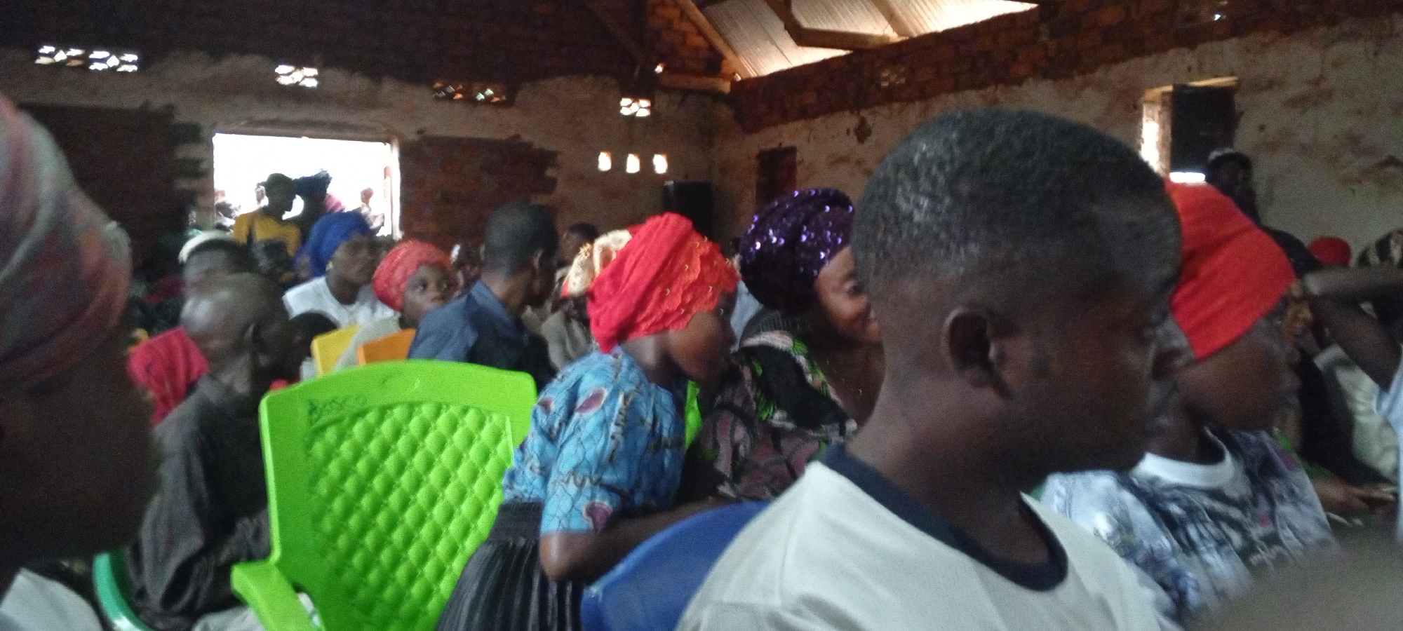 A gathering of people are seated inside what appears to be a church building in Ngalula, Democratic Republic of the Congo. The interior of the building is worn, with unfinished brick walls and small, high windows allowing light to enter. A corrugated metal roof is visible. Most attendees appear to be adults, and several are wearing colorful head coverings. Some are seated on plastic chairs, including a bright green one. The faces of most individuals are visible, including those of children, but many of the faces are obscured.