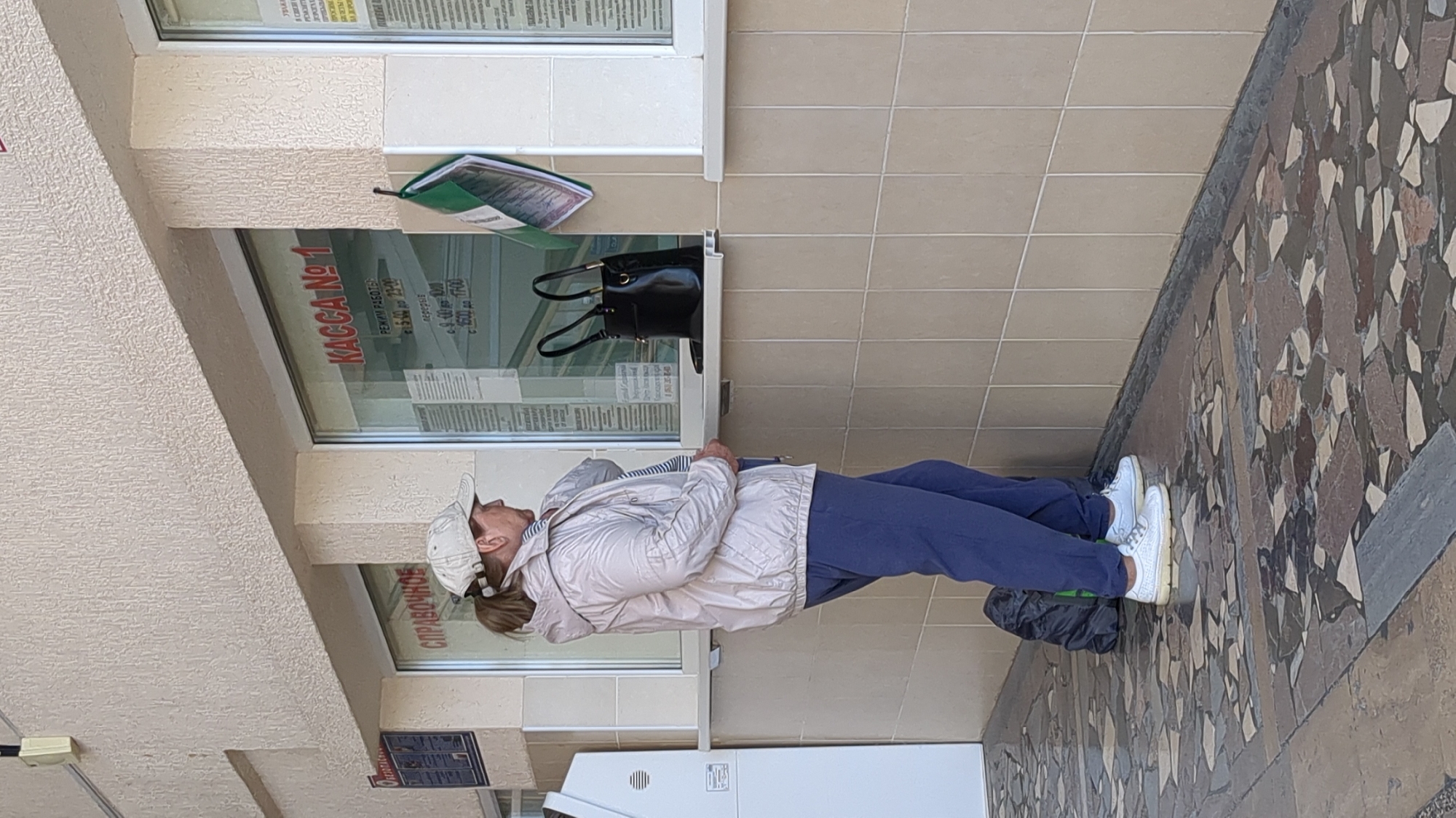 The image captures an outdoor scene in Gelendzhik, Russia, depicting a person standing before a service window of a building. The individual, appearing to be a woman, is dressed in a light-colored, possibly beige or white, hooded jacket over a striped shirt, dark blue sweatpants, and white sneakers. She wears a light-colored baseball cap and stands with her right hand slightly raised, suggesting she is either waiting or interacting at the window.
The building's exterior features a light-beige, textured wall. Two large windows are prominent, displaying various documents and signs behind the glass. On the sill separating the two window panes, a black handbag and a green folder containing papers are placed. Below the window, a white utility unit is partially visible. The ground beneath has two distinct patterns: a section of light beige square tiles closer to the building, and a more distant section featuring a mosaic-like arrangement of brown, grey, and white geometric pieces.
Visible text on the right window prominently displays "KACCA №1" (Cashier No. 1) in red. Below this, "РЕЖИМ РАБОТЫ" (Operating Hours) is detailed as "c 8.00 до 16.00" (from 8:00 to 16:00), with a "Перерыв" (Break) scheduled "c 16.00 до 17.00" (from 16:00 to 17:00). Other smaller text, possibly indicating days of the week like "ПН - СБ" (Monday - Saturday), is also present. On the left window, the word "СПРАВОЧНОЕ" (Inquiry/Information) is clearly visible.
The lighting suggests it is daytime, with clear, bright illumination indicative of a sunny or clear weather day. The scene portrays a routine interaction or waiting period at a service counter in an urban setting.
