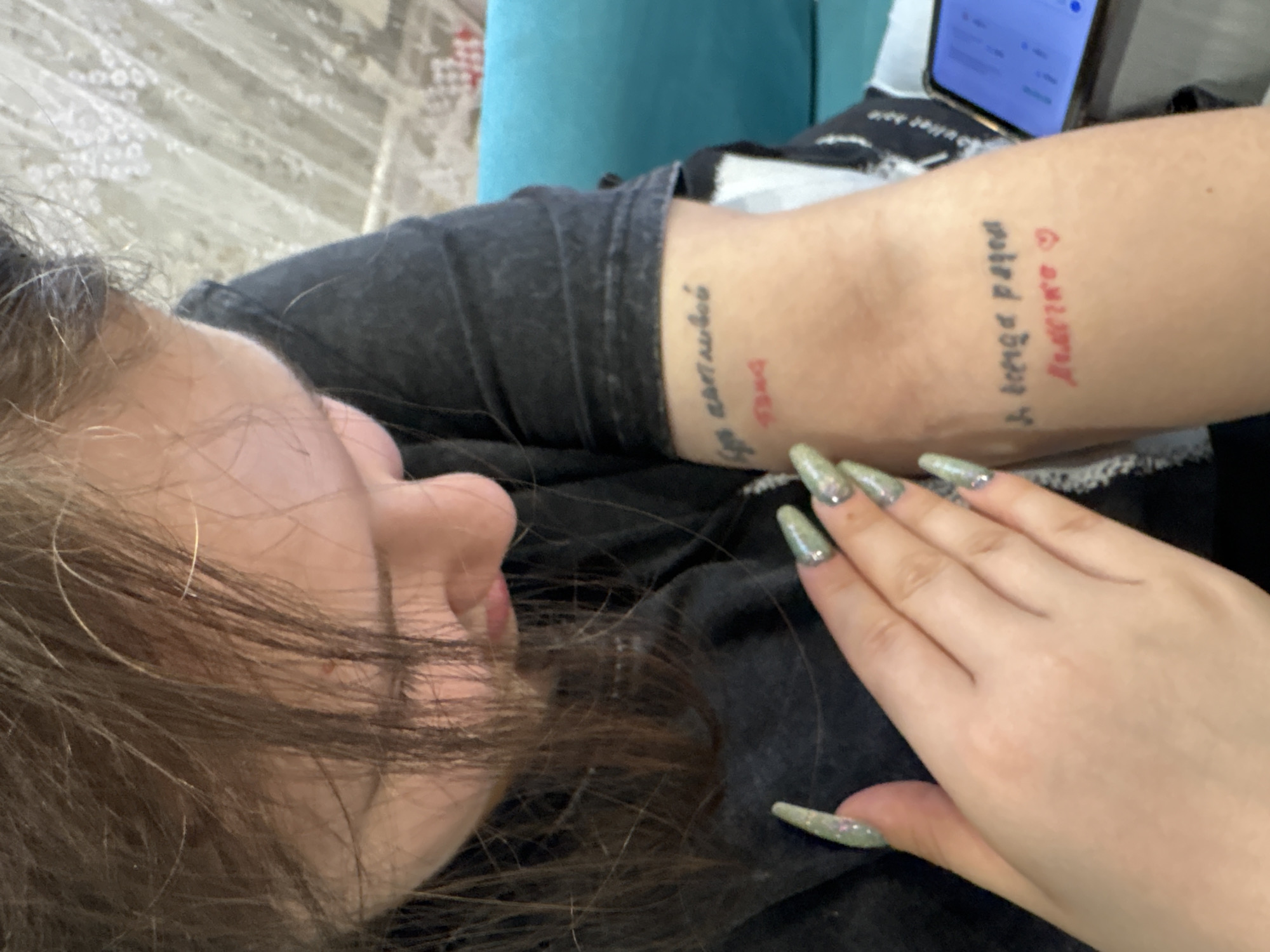 A young person with long brown hair is lying down, facing left, with her right arm bent and resting on her torso. Her hand is prominently displayed, showing long, green nails with glitter accents. Tattoos are visible on both forearms. The tattoos appear to contain both English and Cyrillic script. A mobile phone is partially visible in the upper right corner. The person is wearing a dark gray shirt. The location of Chaikovsky, Russia cannot be confirmed from the image.