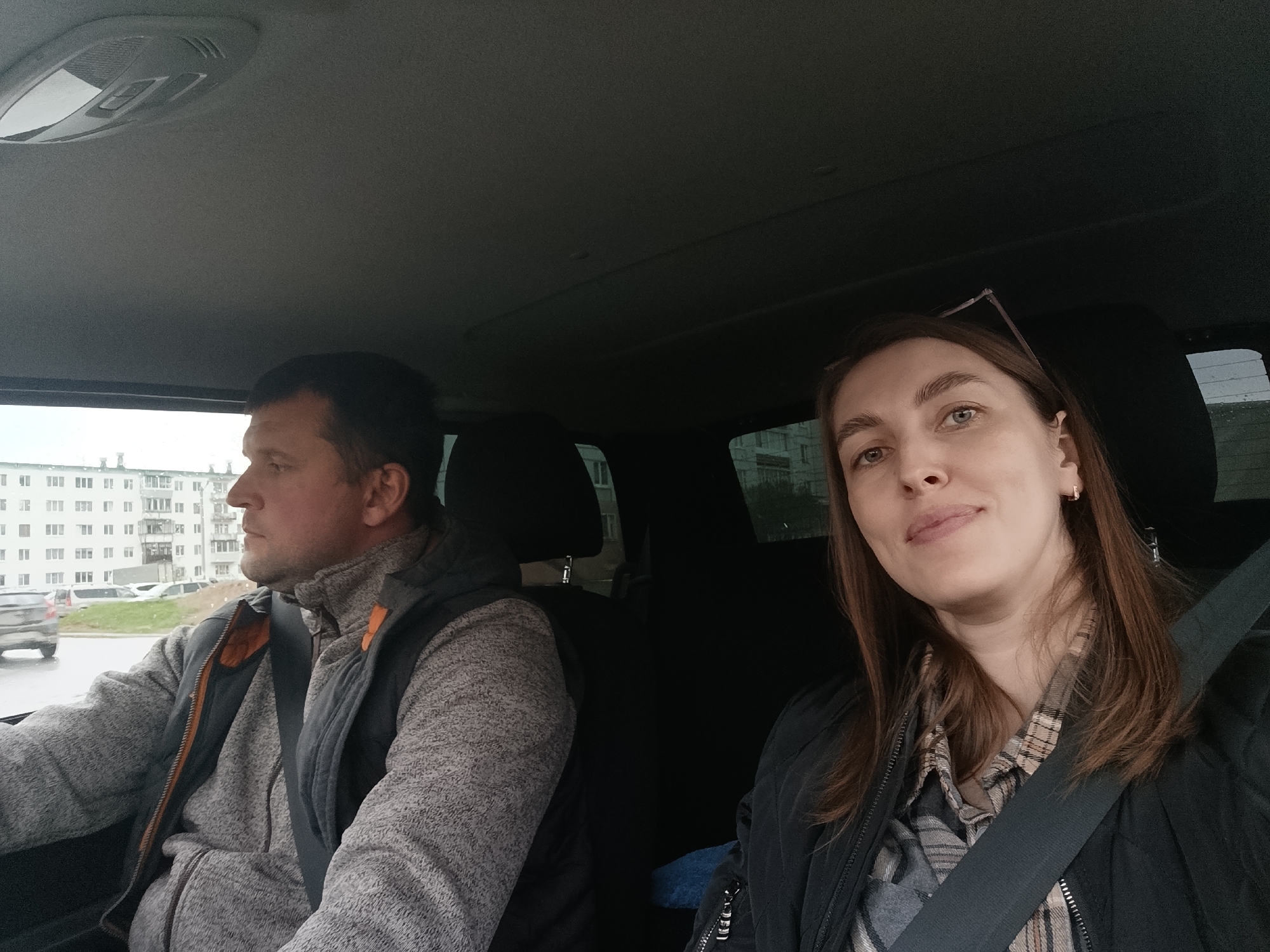 Two people, a man and a woman, are seated in the front seats of a car. Both are wearing seatbelts. The man, on the left, is looking out of the window. He is wearing a gray zip-up sweater and a dark vest. The woman, on the right, is looking at the camera. She is wearing a plaid shirt and a black jacket, and has a pair of glasses resting on her head. The car is moving along a street in Chaikovsky, Russia. Apartment buildings are visible outside the window. Other cars are visible. The weather appears overcast.