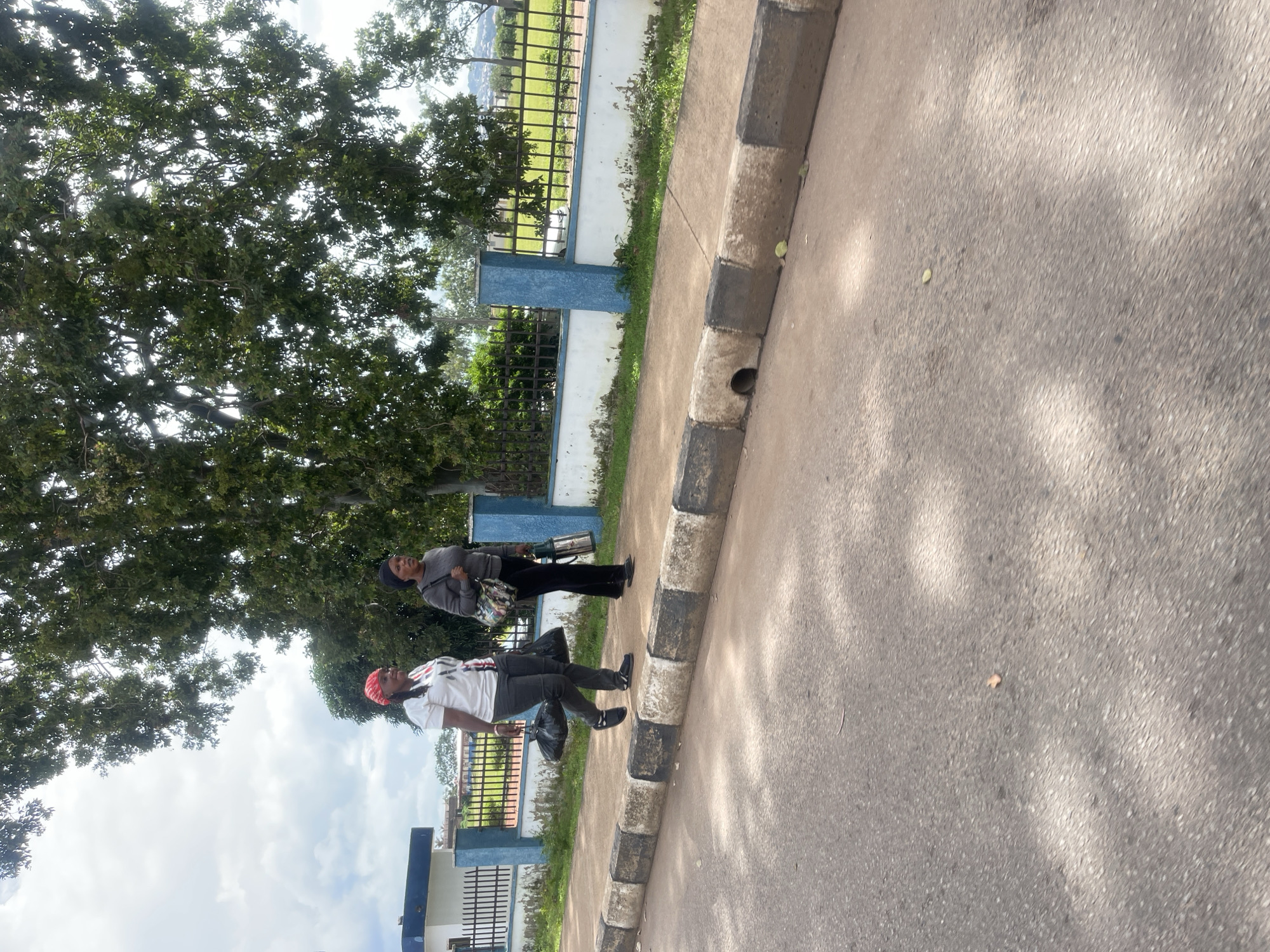 Two women are walking on a sidewalk beside a road in Gura Top, Nigeria. The woman on the left wears a white t-shirt with red and black accents, dark pants, and a red headscarf. She carries a black bag in her left hand and another in her right. The woman on the right wears a grey long-sleeved shirt, black pants, and a black headscarf. She carries a multi-colored bag on her left shoulder and a silver thermos in her right hand. They are walking from left to right in the frame. Behind them is a white and blue fence, a building, and a large tree. The road in front of them is asphalt with a curb that has black and white stripes. The weather appears overcast.