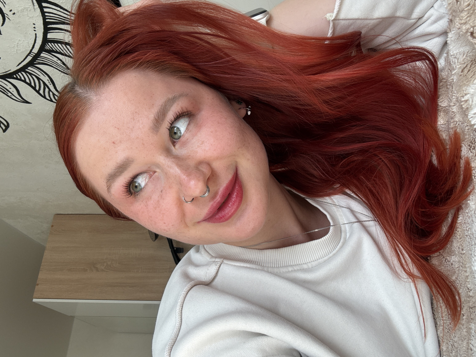 A high-angle portrait captures a young woman with vibrant, long red hair, seemingly taken while she is lying down or lounging indoors. She gazes playfully to the right of the frame with a gentle smile. Her fair skin is adorned with numerous freckles across her nose and cheeks, and her striking green eyes are enhanced with light mascara.
She features a double hoop nose ring and small silver ear piercings on her visible right ear. Her lips are a soft, rosy pink. Her hair, which appears to be dyed a rich auburn or red shade, flows around her, revealing slightly darker roots near her scalp.
The woman is dressed casually in a cream or off-white crewneck sweatshirt, over which a thin silver chain necklace is visible. In the background, a white wall is partially covered by a black and white tapestry featuring a stylized sun or mandala design. Her hair rests on a soft, light-colored textured fabric, possibly a blanket. A light wooden surface, like a shelf or table, can be seen in the lower right portion of the image. The lighting suggests a bright indoor setting during the daytime. The overall impression is relaxed and cheerful. No visible text is present in the image. The provided location context for this image is Rostov-on-Don, Russia.
