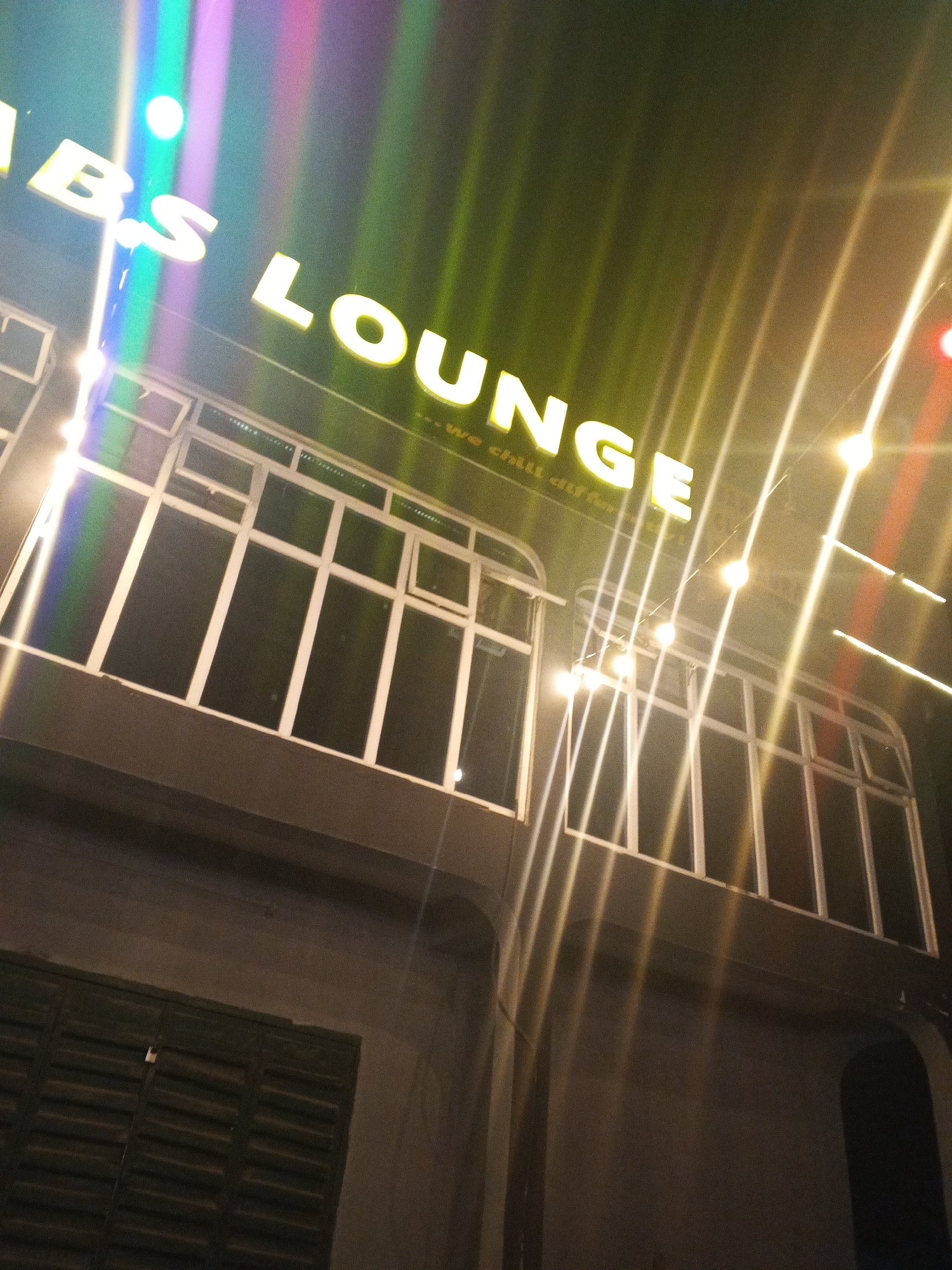 The image depicts the exterior of a commercial building at night in Lokoja, Nigeria, likely a lounge or bar. The scene is dominated by a bright neon sign in white capital letters that reads "BS LOUNGE," with the initial letters of the first word (presumably "VIBES") cropped out on the left. Below "LOUNGE," smaller, faint text is partially visible, appearing to spell "...we chill different."
The building features a dark facade, possibly dark grey or black, which contrasts sharply with the bright lighting. A prominent band of windows with distinct white frames runs across the upper floor, suggesting an open or inviting interior. Numerous string lights, emitting warm yellow light, are strung horizontally across the building, creating striking lens flares and light streaks that cut diagonally through the frame. Colorful light effects, including shades of purple, blue, and green, are also visible, particularly around the "BS" section of the sign, indicating dynamic ambient lighting.
The lower part of the building shows dark walls. On the bottom left, a dark, slatted structure, possibly a closed shutter or gate, is present. To the right, a dark arched entrance or window is visible. The photograph is taken from a low angle, looking upwards, emphasizing the illuminated signage and the architectural details against the dark night sky. No people or explicit activities are visible, focusing solely on the establishment's facade and its nighttime illumination.