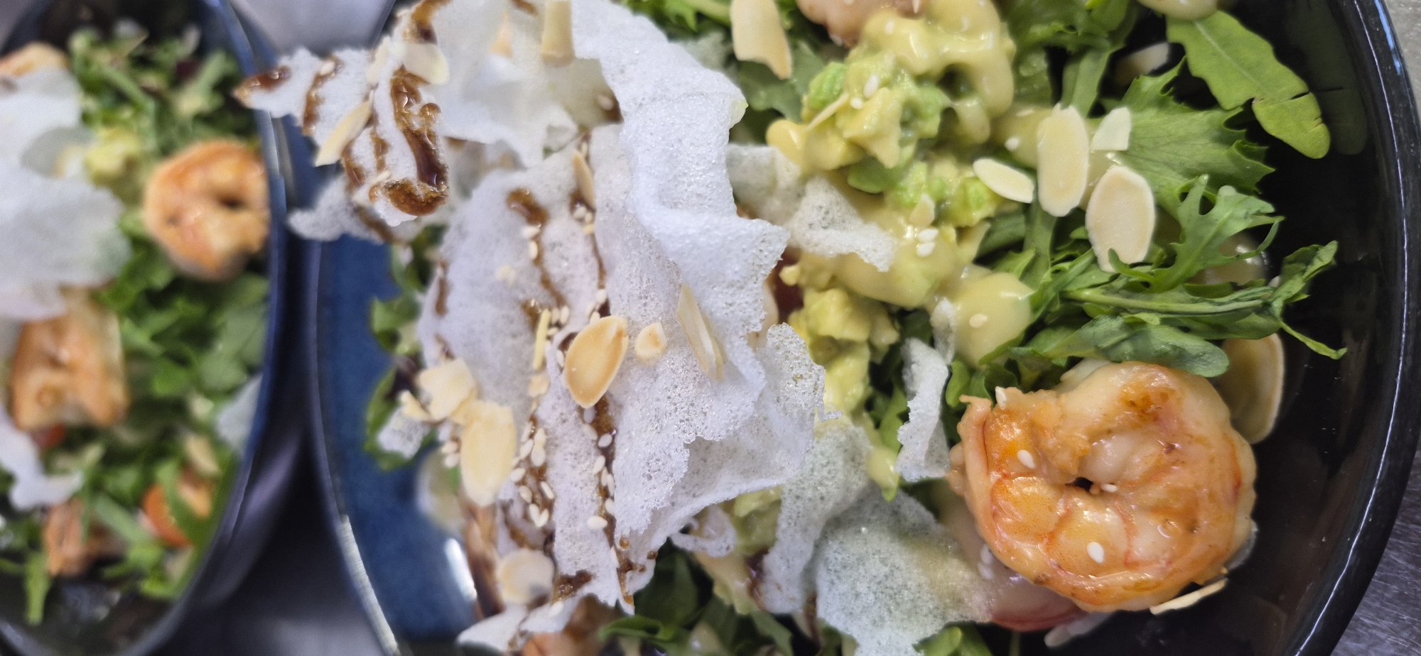 This is a close-up, overhead shot of a shrimp salad presented in a dark blue bowl. The salad contains leafy greens, diced avocado, sliced almonds, and sesame seeds, all drizzled with a creamy dressing and a dark sauce. One large, cooked shrimp is prominently displayed in the lower left corner of the bowl. In the background, a second, out-of-focus bowl of a similar salad is visible. The image appears to have been taken indoors, likely in a restaurant setting in Saint Petersburg, Russia, based on the context provided. There are no people visible in the image.